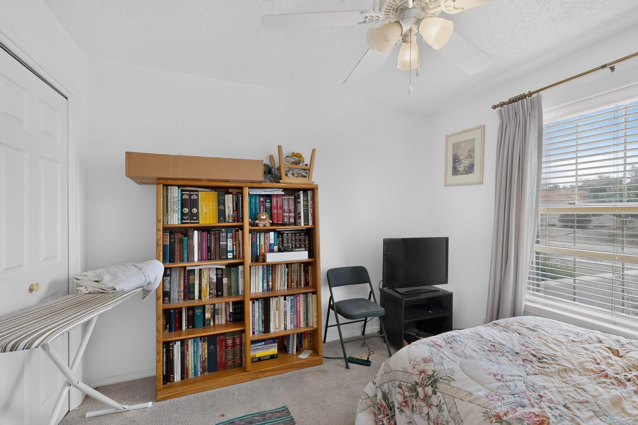 Bedroom with a book-filled wooden bookcase, floral bed, and television beside a large curtained window.