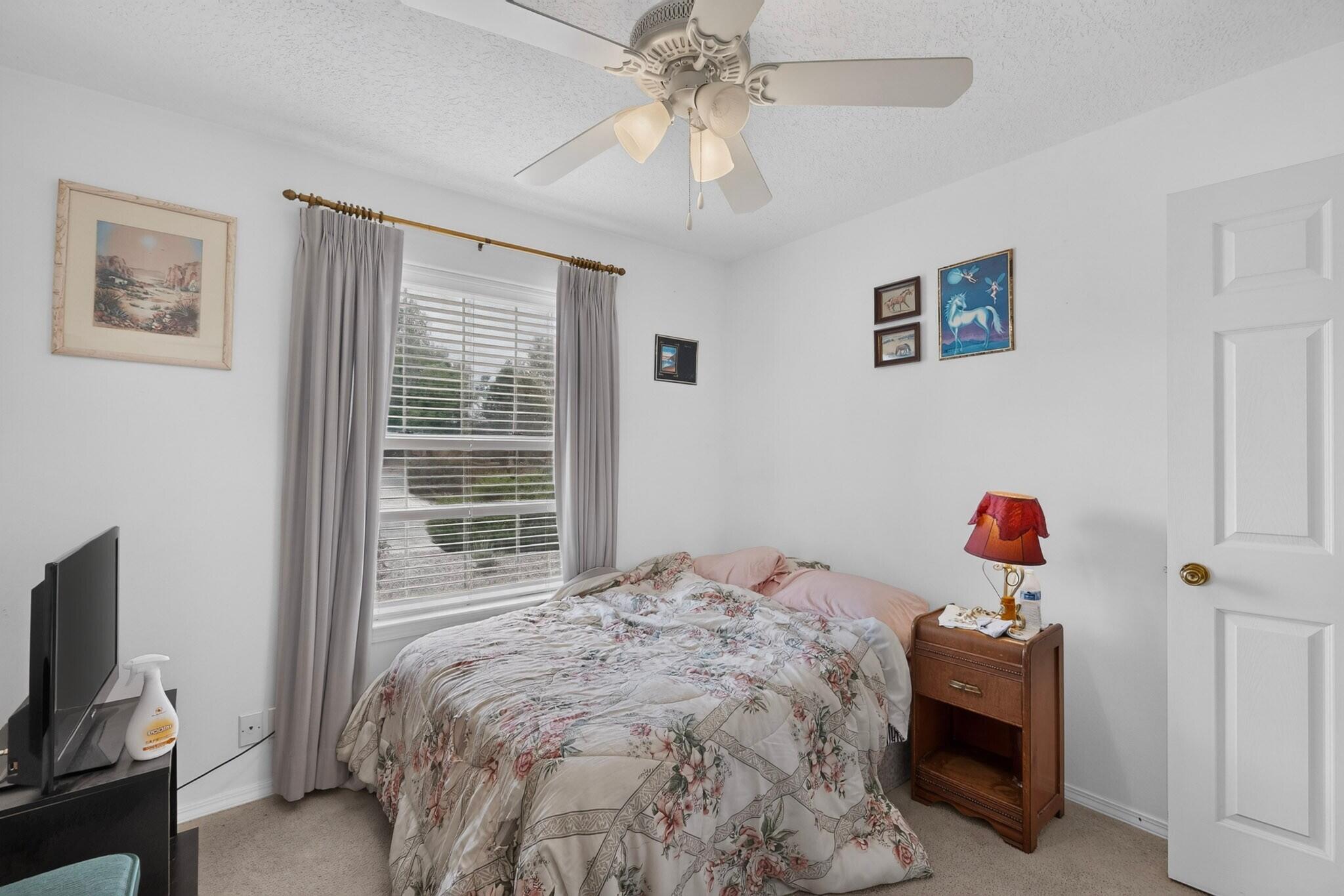 Bedroom with a floral bedspread, curtained window, ceiling fan, and wooden bedside table.