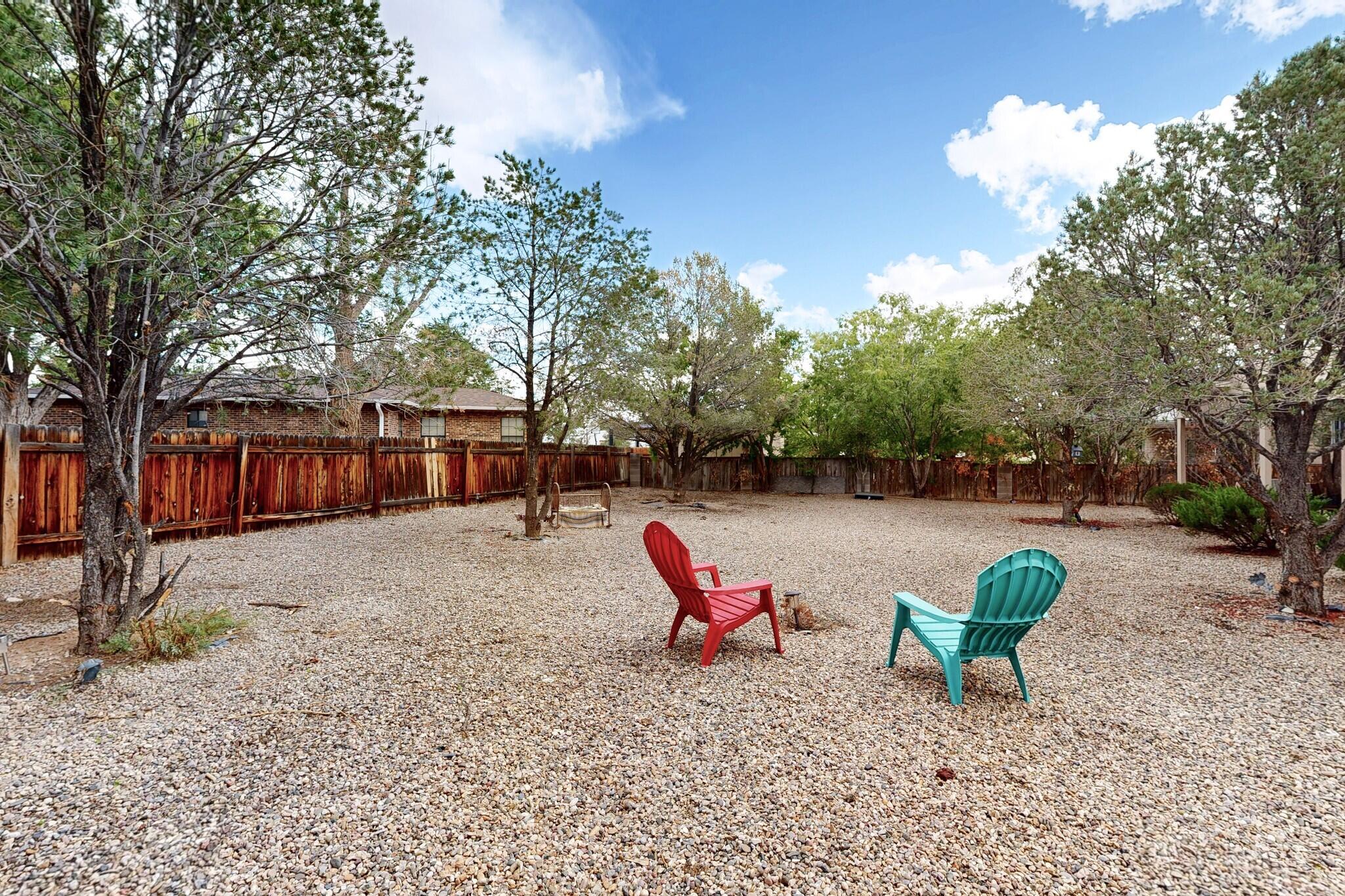 Gravel backyard with mature trees and two colorful Adirondack chairs inside a wooden fence.