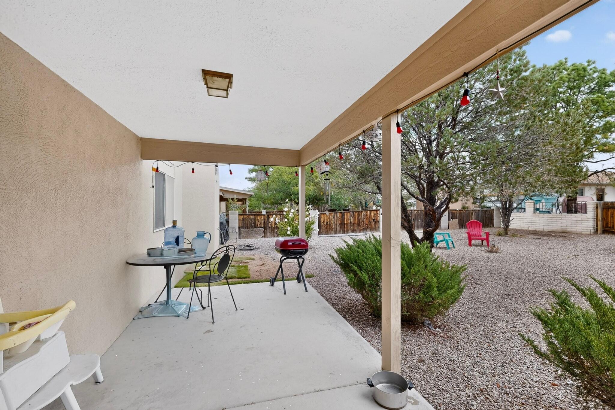 Covered patio with a red grill and string lights beside a gravel backyard with trees and colorful chairs.