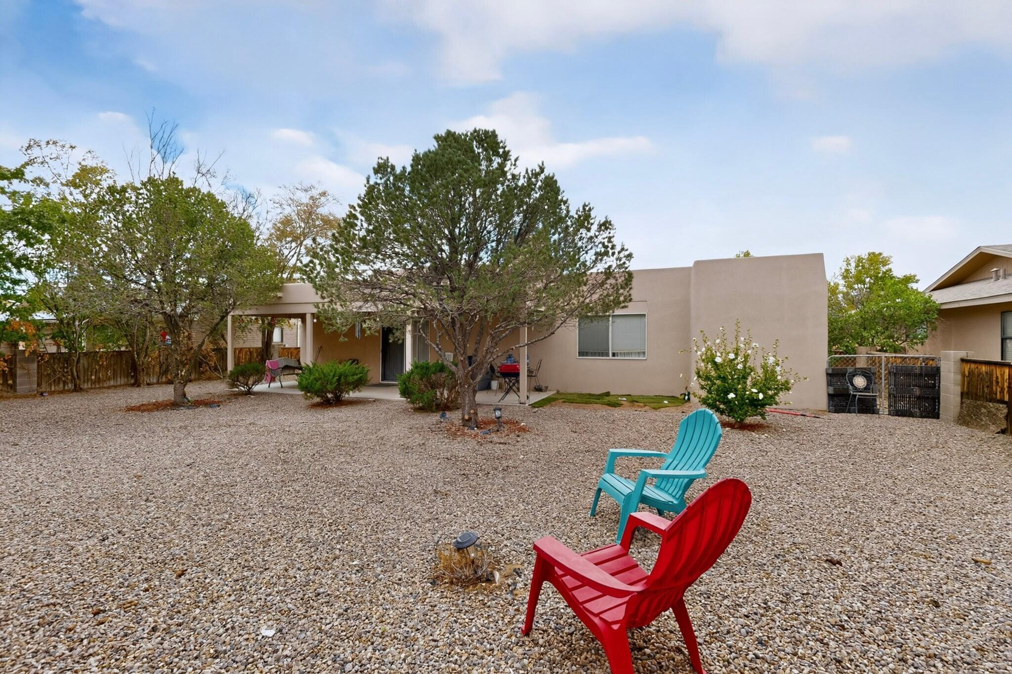 Backyard with a gravel yard, covered patio, trees, and red and turquoise chairs behind a beige stucco home.