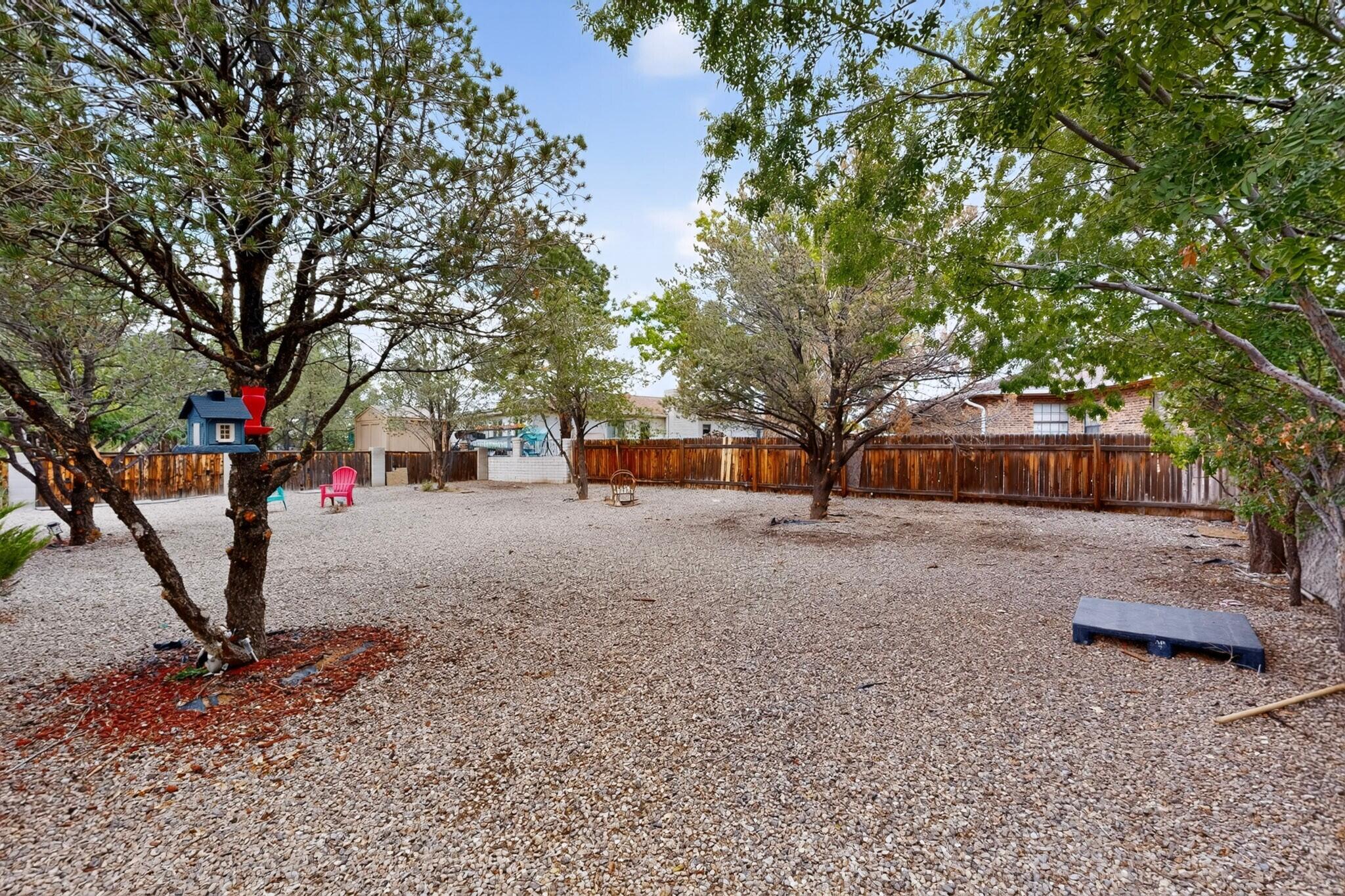 Gravel backyard with mature trees, a wooden fence, birdhouse, and colorful chairs.