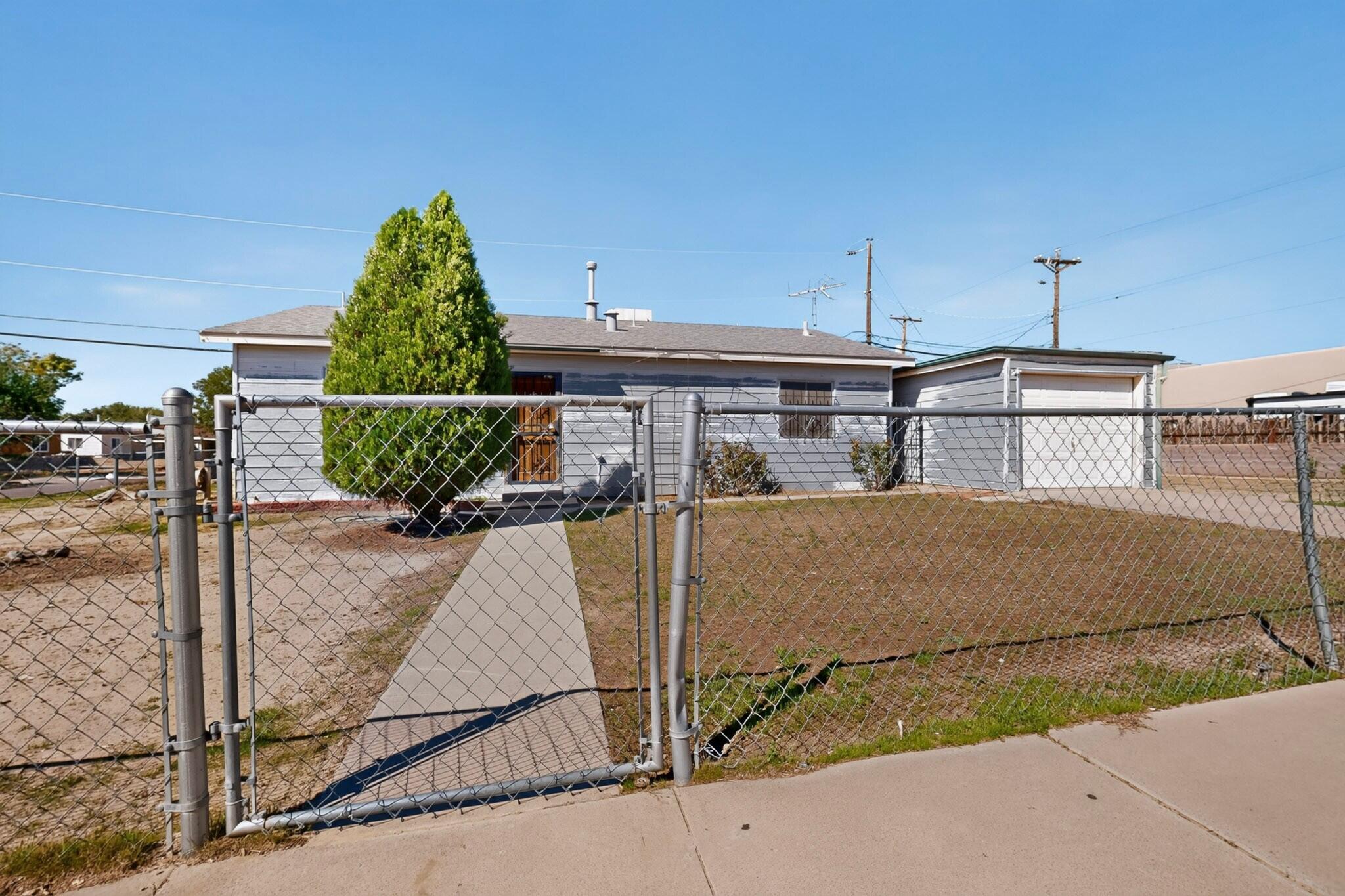Gray single-story house behind a chain-link fence, with a concrete walkway and attached garage.