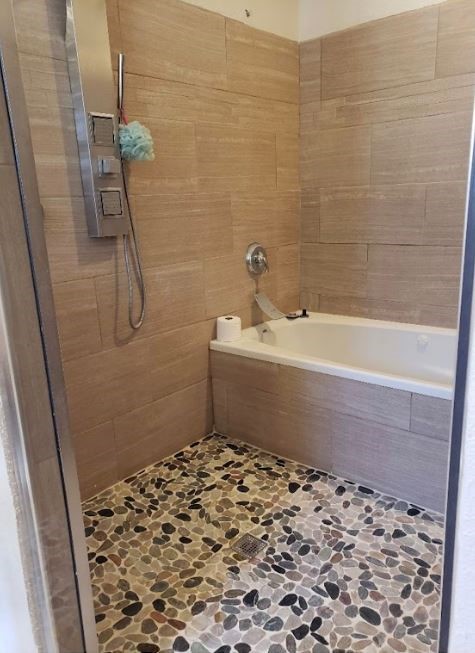 Bathroom with a built-in tub, handheld shower, tan tile walls, and pebble-stone flooring.