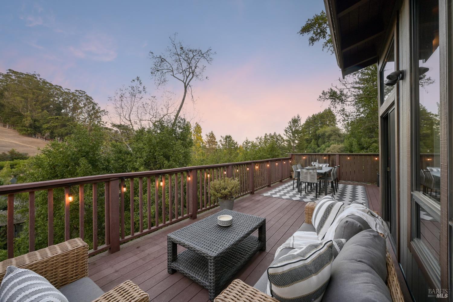 Furnished deck overlooking a wooded hillside, with wicker seating and string lights at dusk.