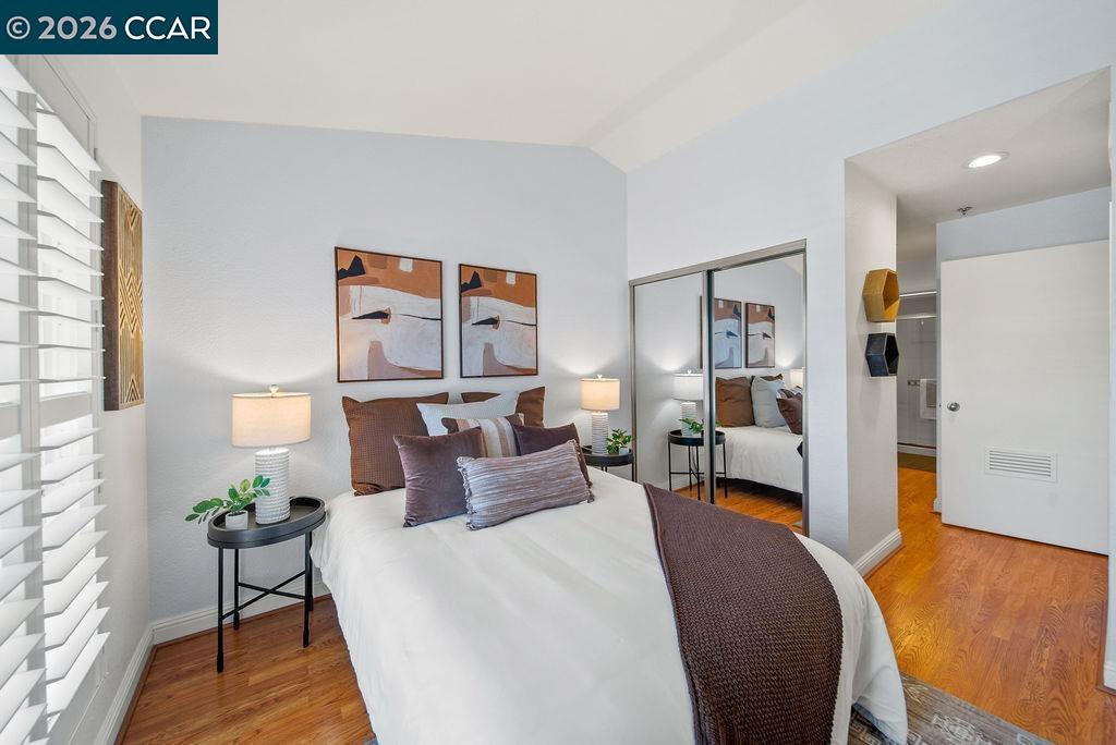 Staged bedroom with mirrored closet doors, hardwood floors, and a white bed with brown accents.