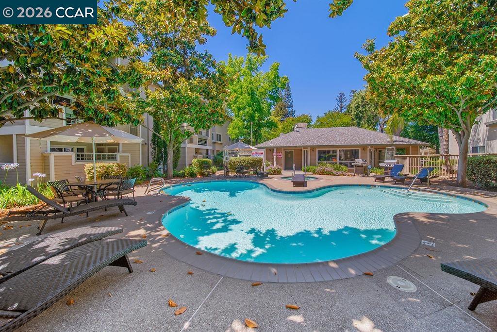 Outdoor swimming pool surrounded by apartment buildings, patio seating, and mature trees.