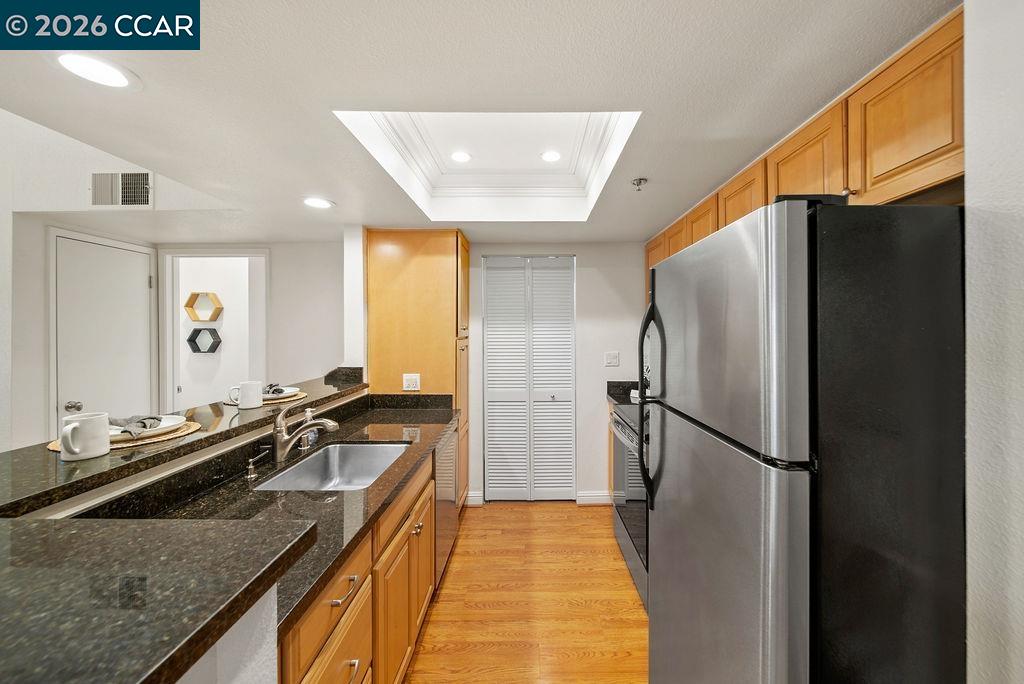 Galley kitchen with granite counters, warm wood cabinetry, stainless steel refrigerator, and recessed tray ceiling.