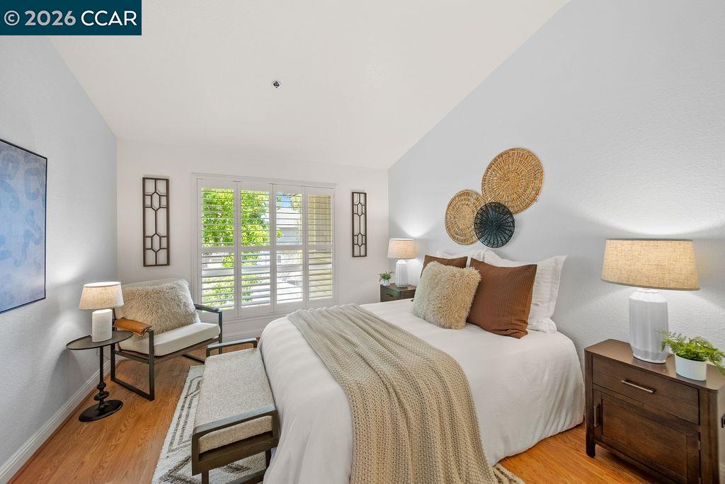 Staged bedroom with a white bed, woven wall decor, and shuttered windows overlooking greenery.
