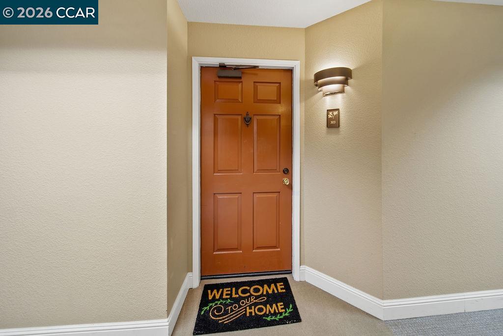 Unit 303 entryway with a reddish-brown paneled door, lit wall sconce, and welcome mat.