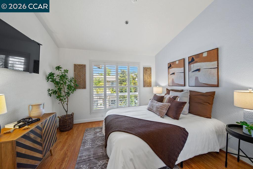 Staged bedroom with a white bed, brown accents, hardwood floors, and shuttered windows.