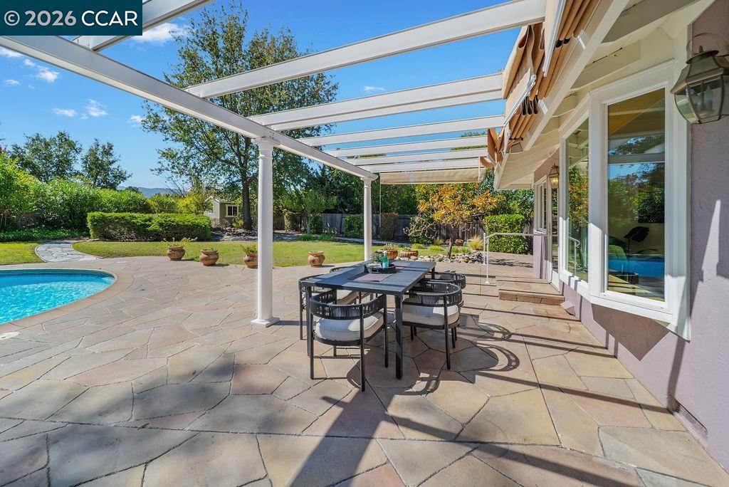 Stone-paved backyard patio with outdoor dining set under a pergola beside a swimming pool.