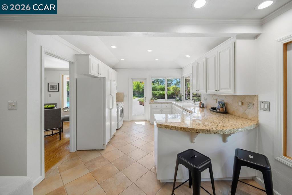 White kitchen with a curved granite breakfast bar, two black stools, and windows overlooking greenery.