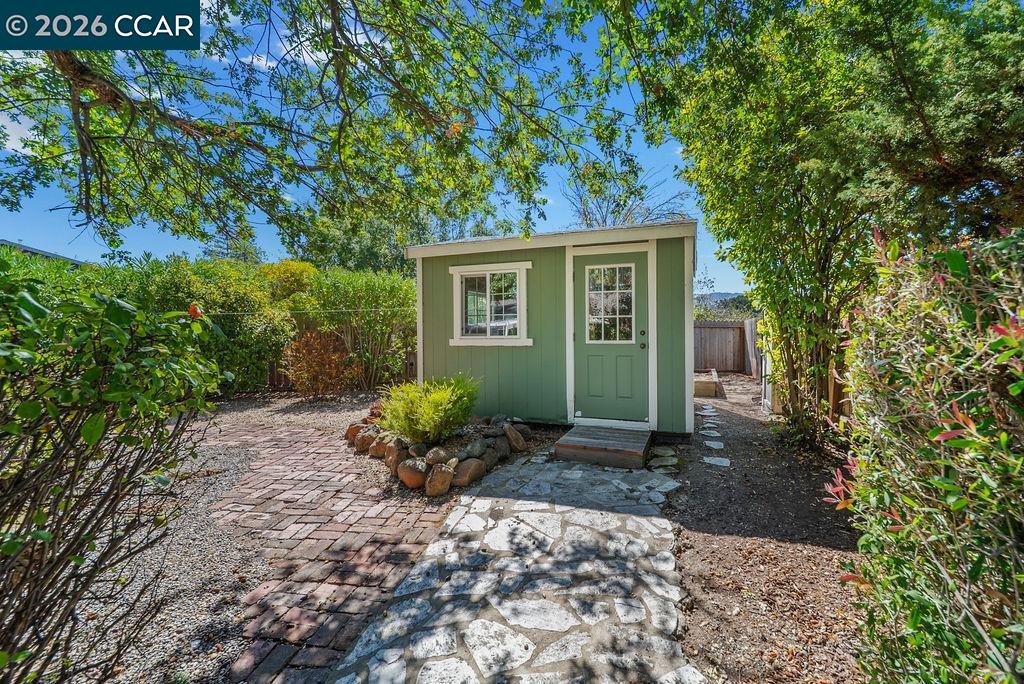 Green garden shed with white trim at the end of a stone path in a leafy backyard.