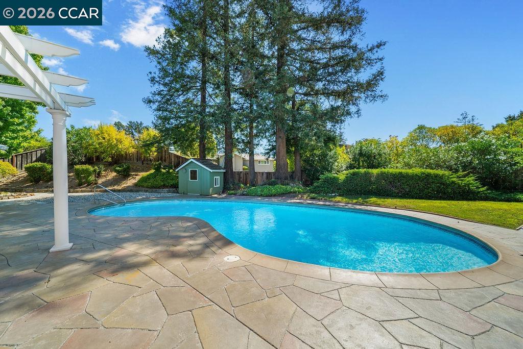 Backyard swimming pool with a flagstone patio, white pergola, and tall trees.