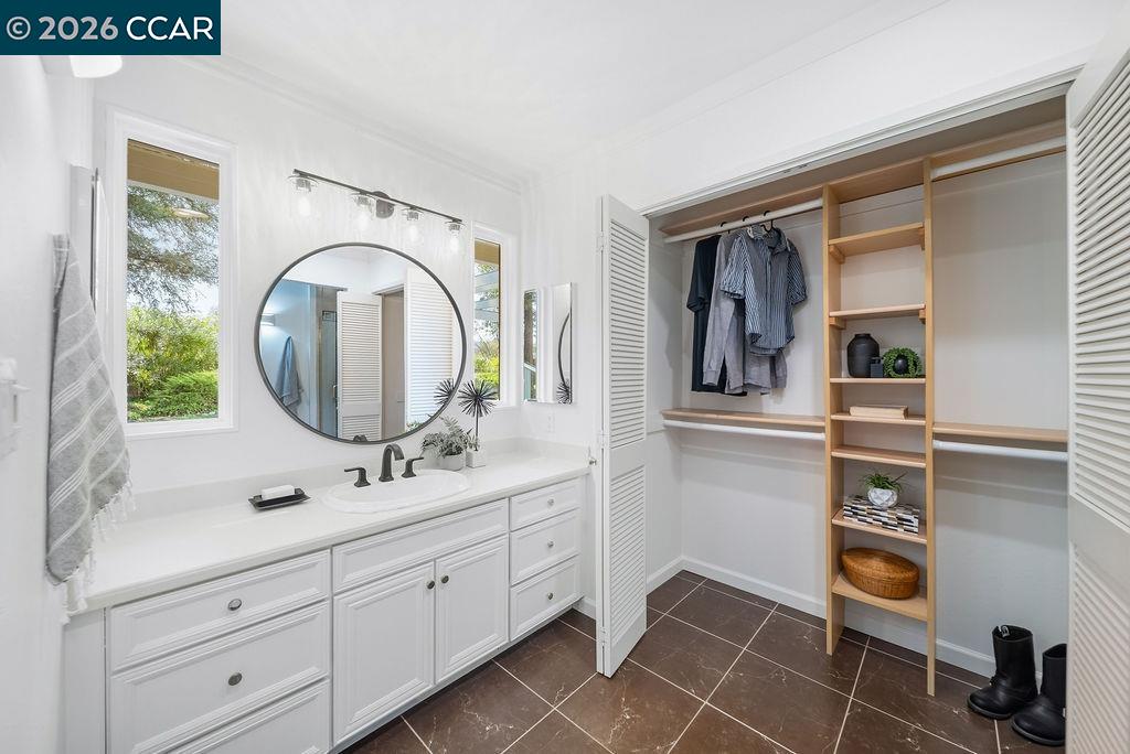 Bathroom with a long white vanity and round mirror opening into a walk-in closet with wood shelving.