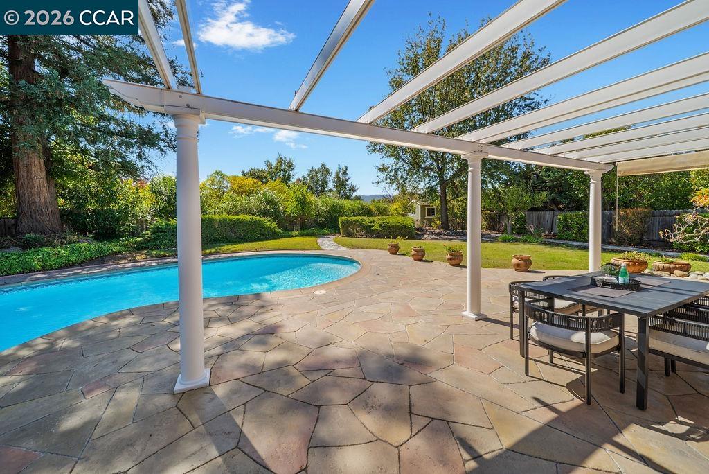 Backyard pool and stone patio beneath a white pergola, with an outdoor dining table.