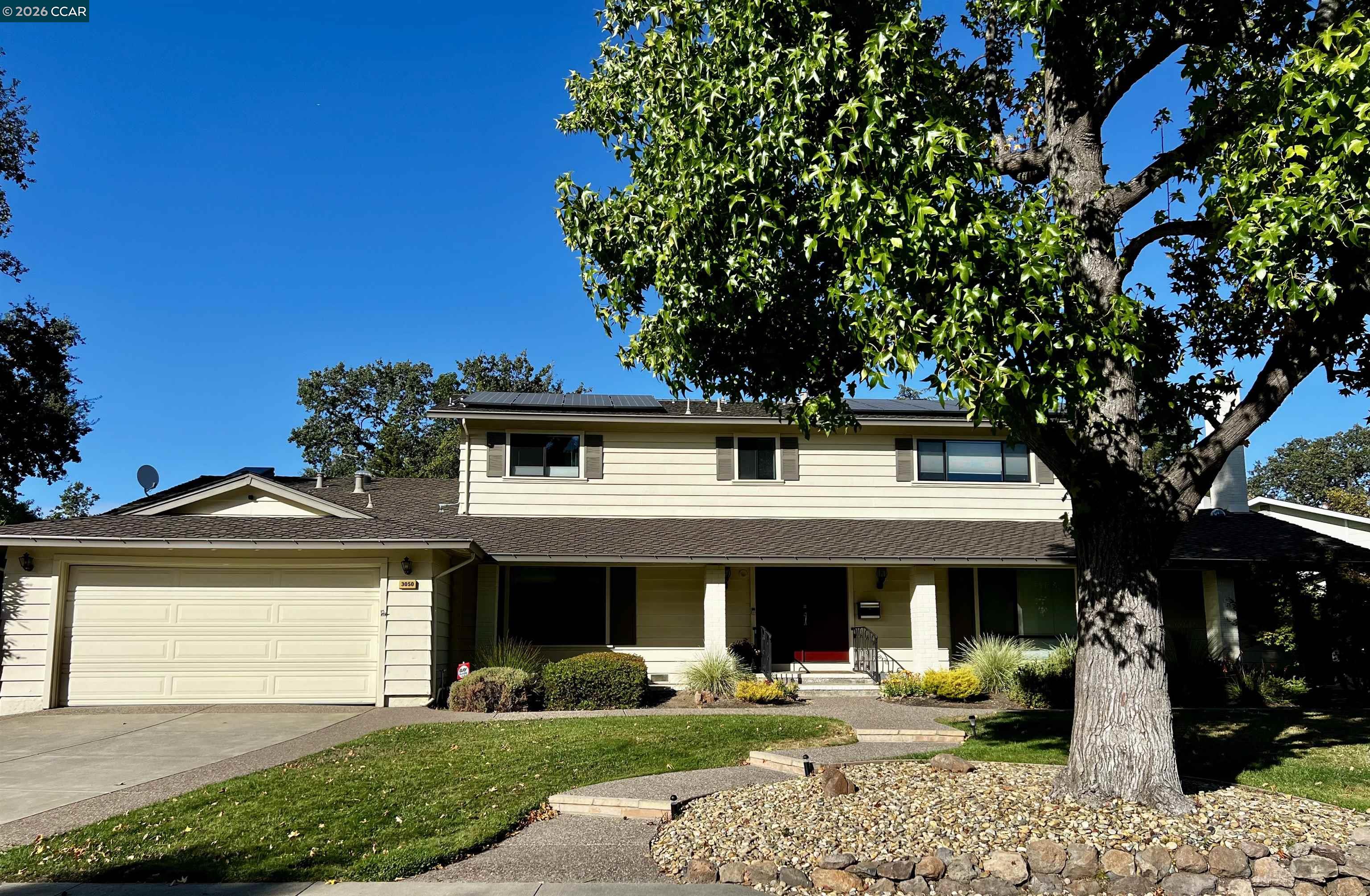 Two-story cream house with solar panels, a wide garage, and a large tree in the front yard.