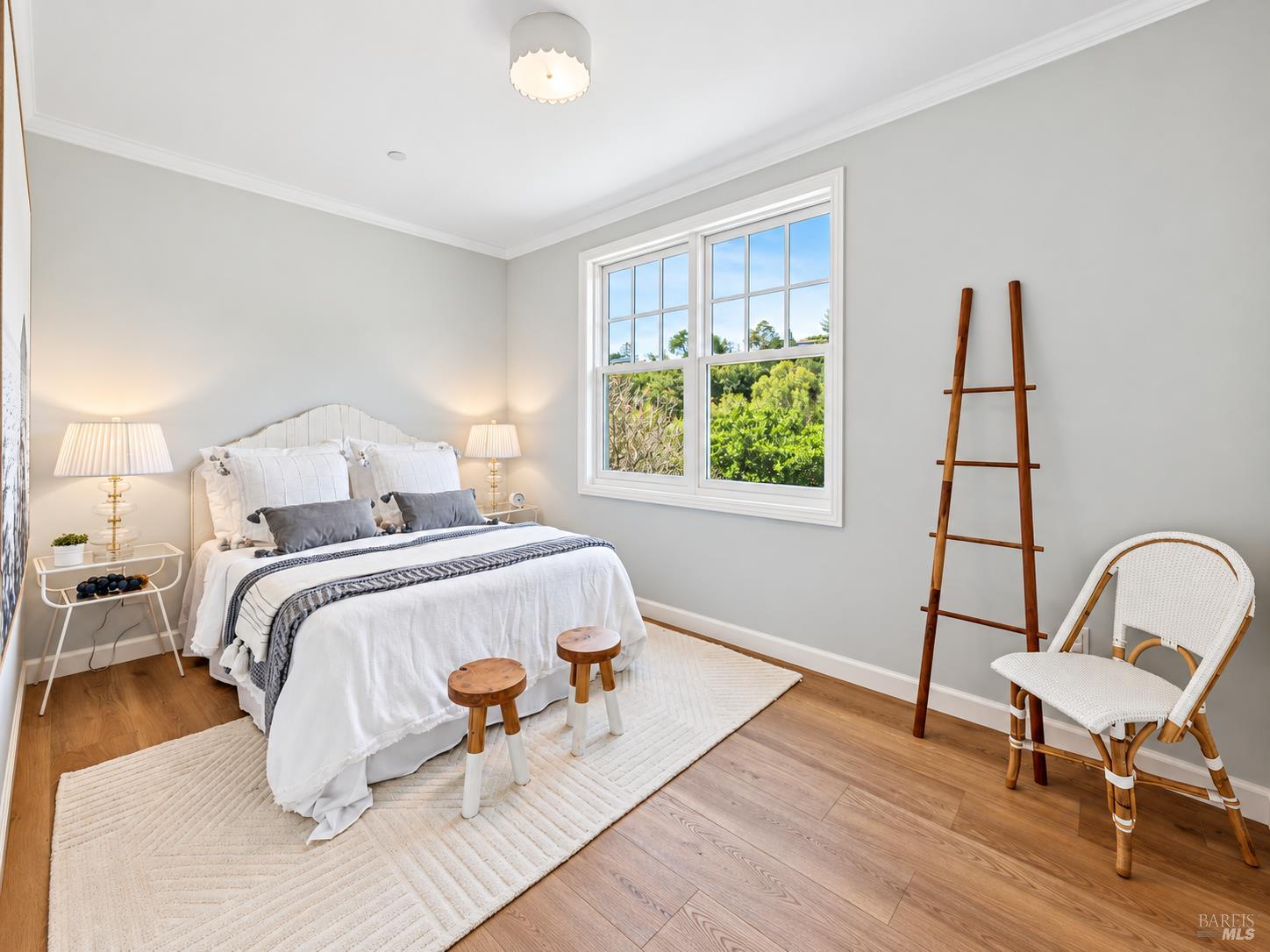 Staged bedroom with a white bed, large window overlooking greenery, and warm wood floors.