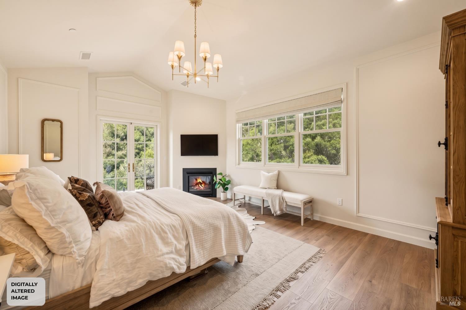 Staged bedroom with a large bed, fireplace, and windows overlooking trees.
