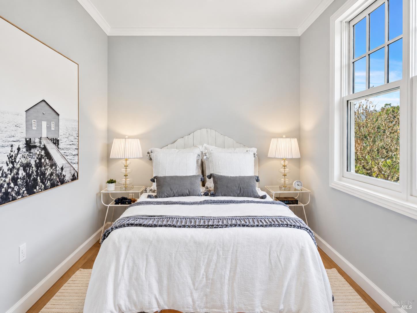 Staged bedroom with a white bed, matching bedside lamps, and a tall window overlooking trees.
