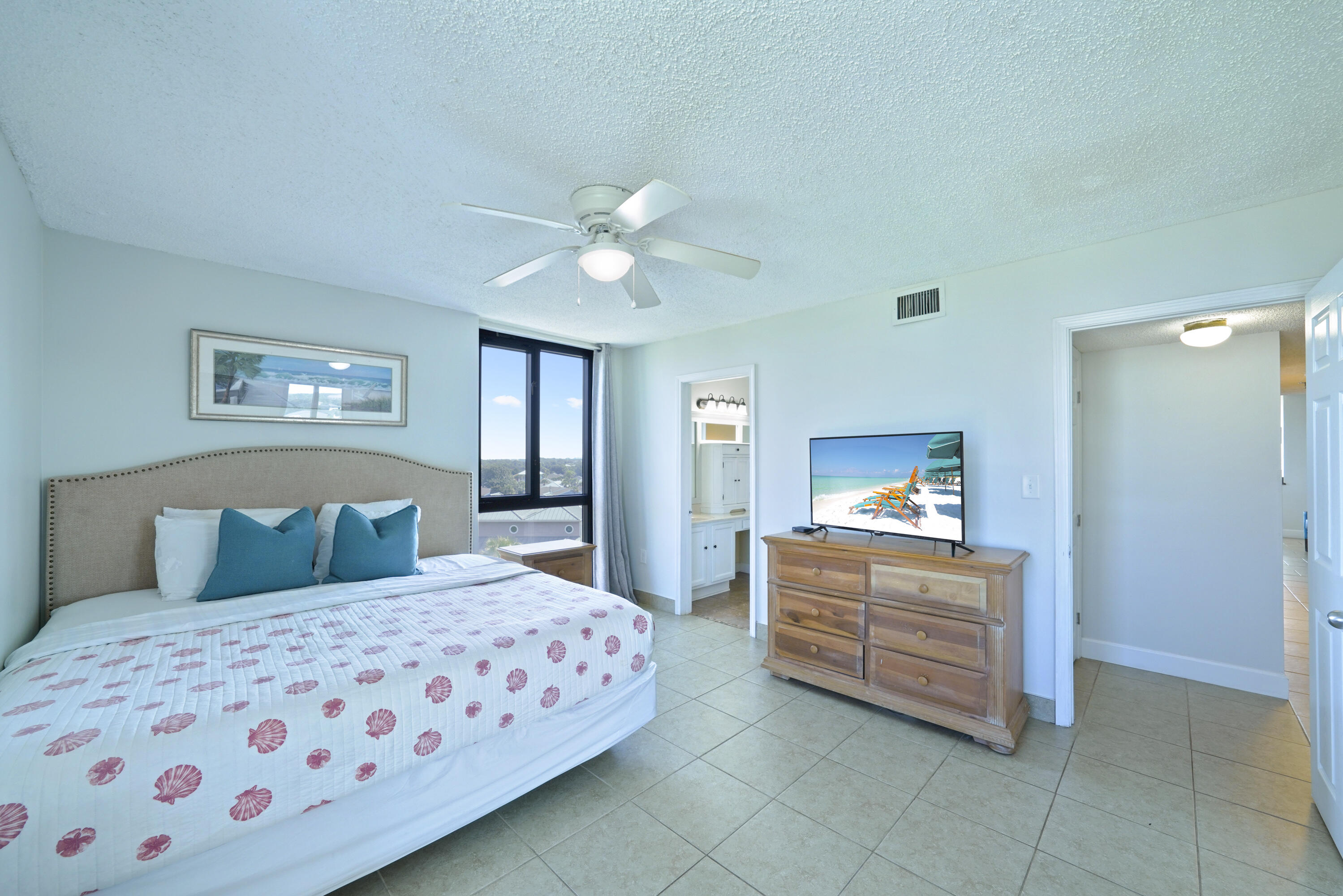 Furnished bedroom with a shell-patterned bedspread, teal pillows, and a wooden dresser beneath a television.