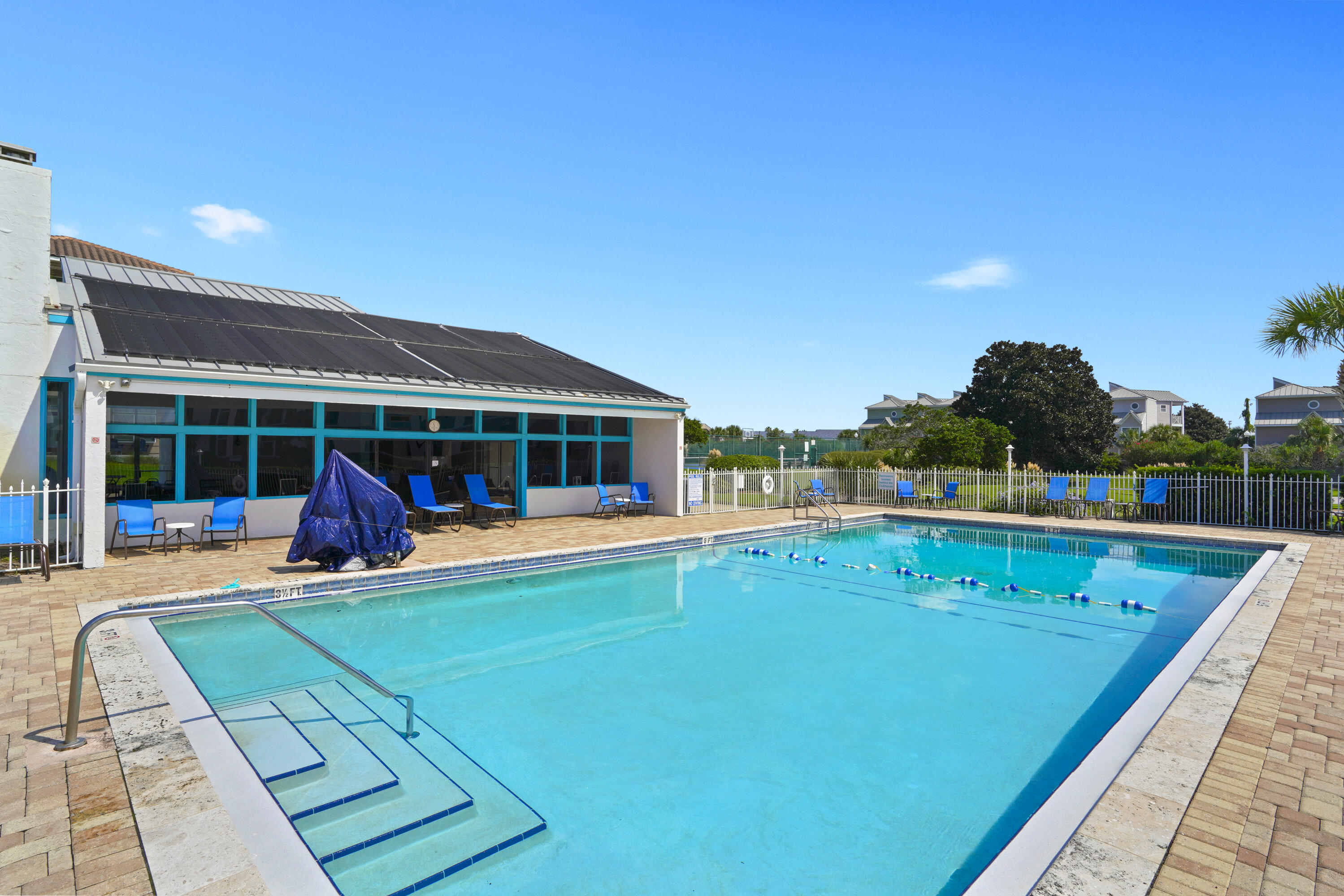 Outdoor pool with wide entry steps, a fenced paver deck, and lounge chairs beside a clubhouse.