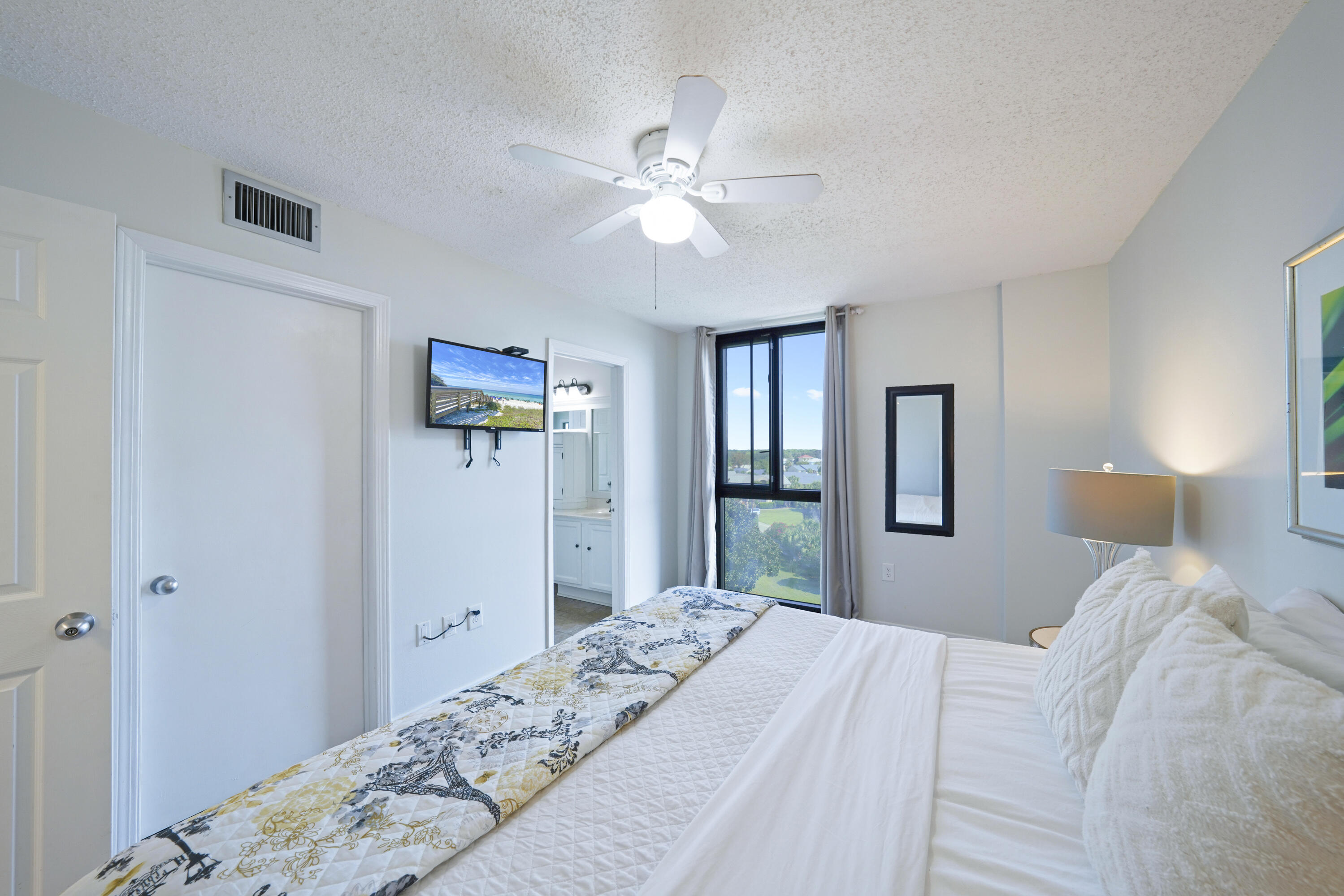 Furnished bedroom with a patterned quilt, ceiling fan, and large window overlooking greenery.