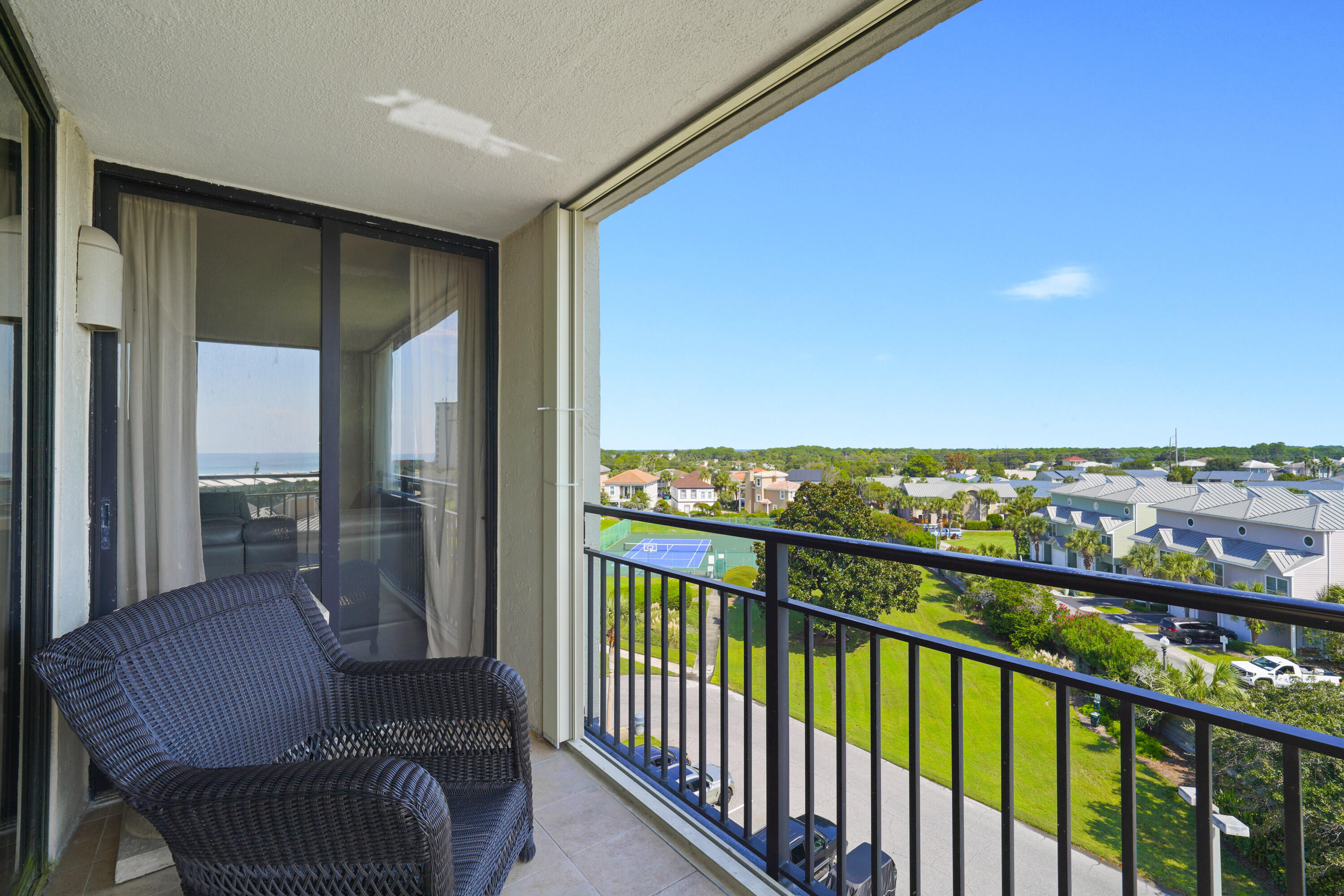 Condo balcony with wicker chairs overlooks a tennis court, green grounds, and nearby homes.