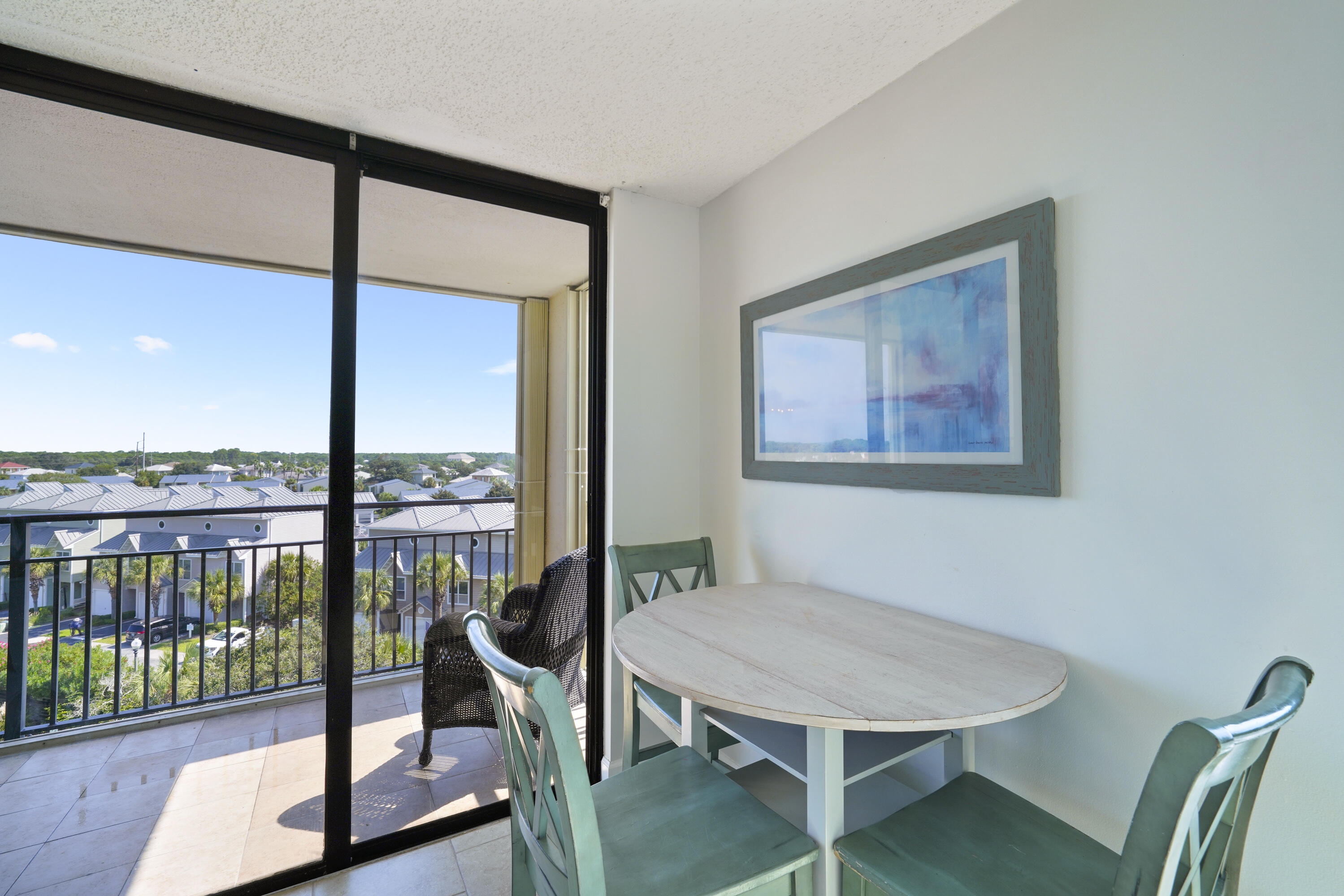 Condominium dining area with a round table and green chairs beside a balcony overlooking nearby homes.