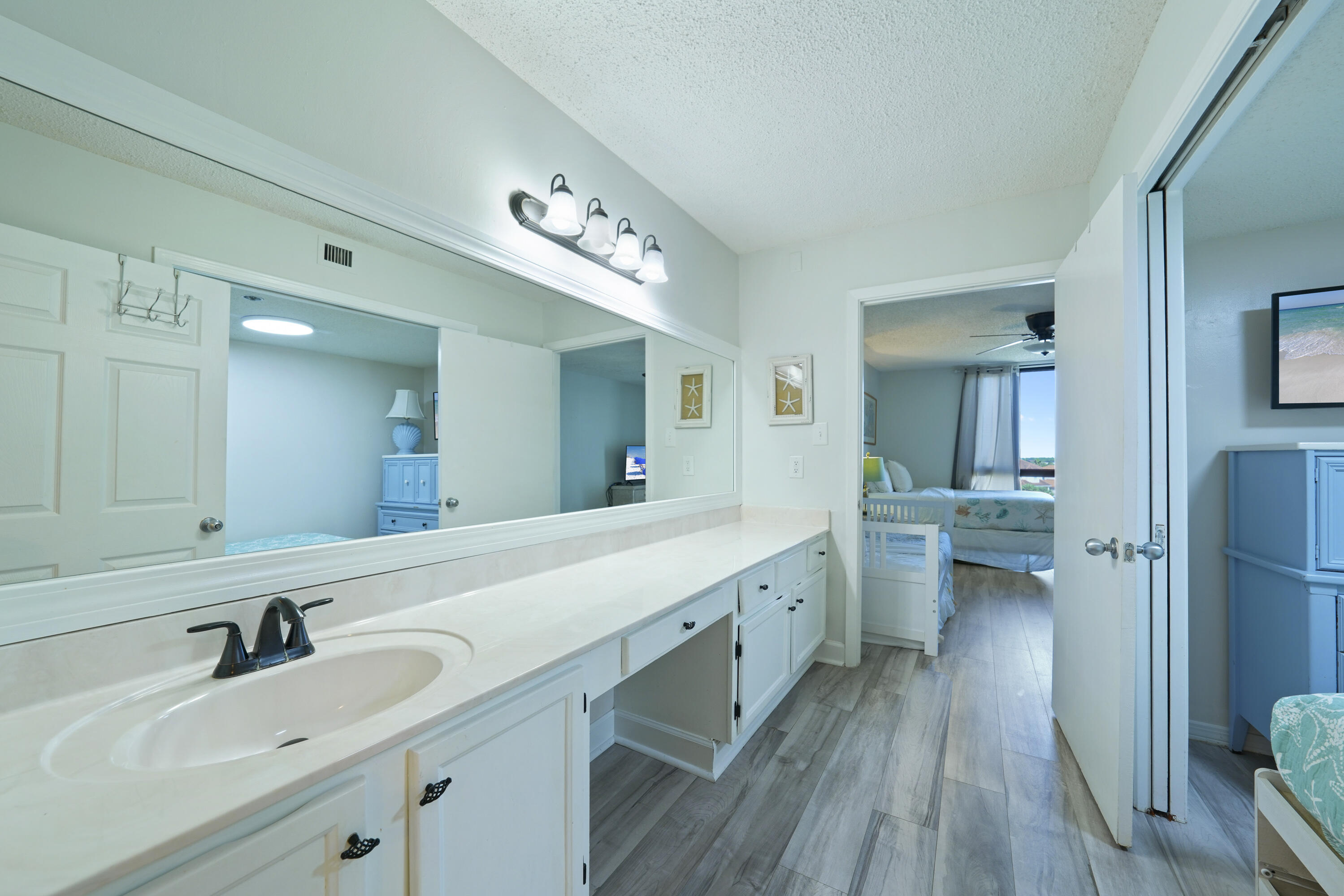 Bathroom vanity with a long white counter and large mirror; an open doorway reveals a blue-accented bedroom.
