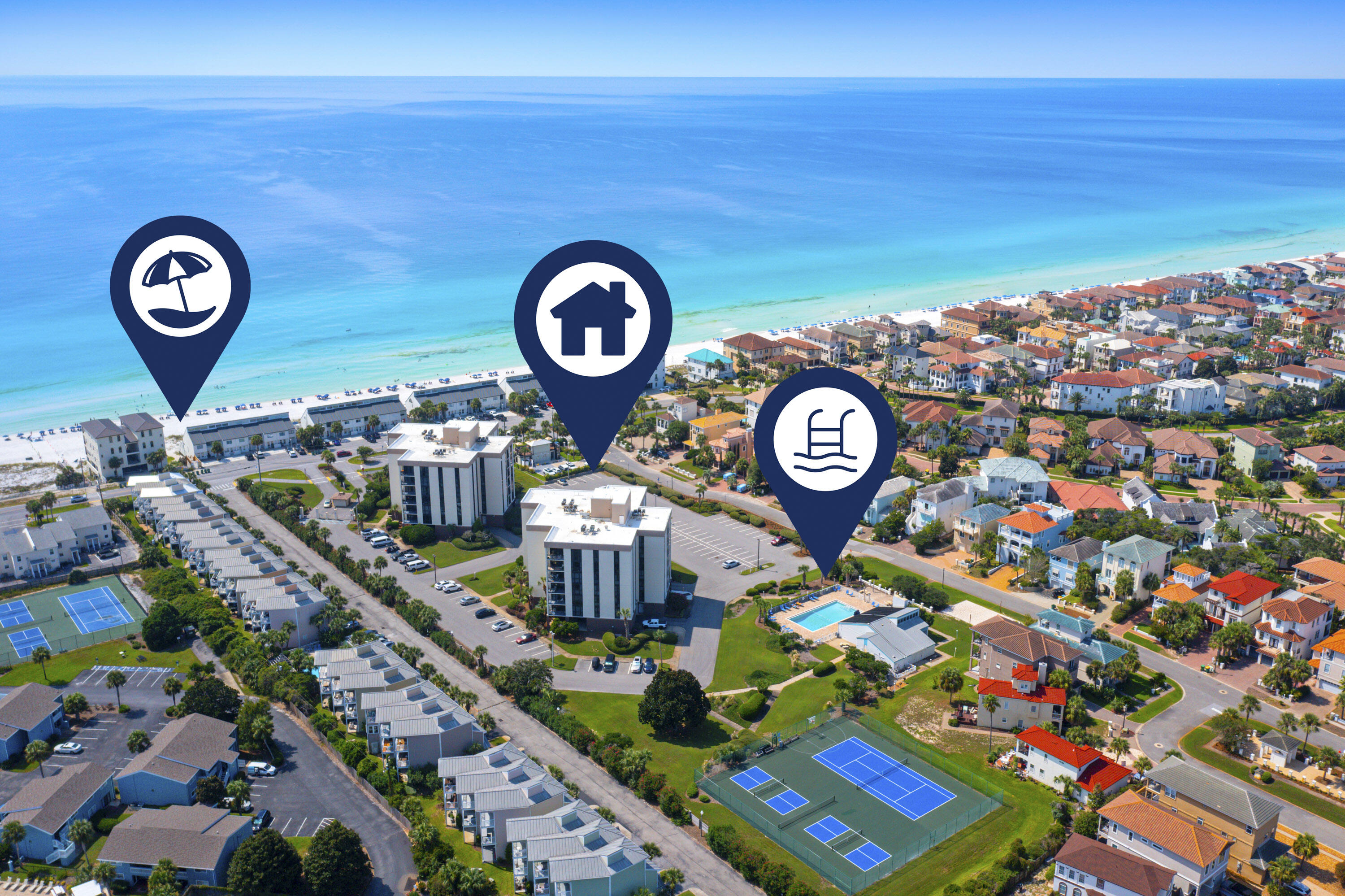 Aerial coastal view of beachside condos, homes, a pool, and tennis courts beside turquoise water.