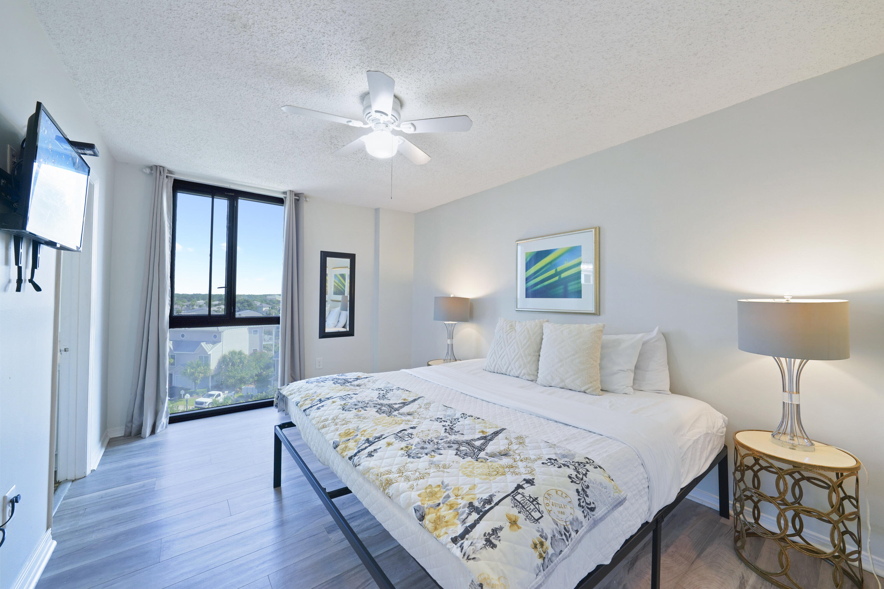 Bright bedroom with a white bed, patterned quilt, and tall window overlooking nearby homes.