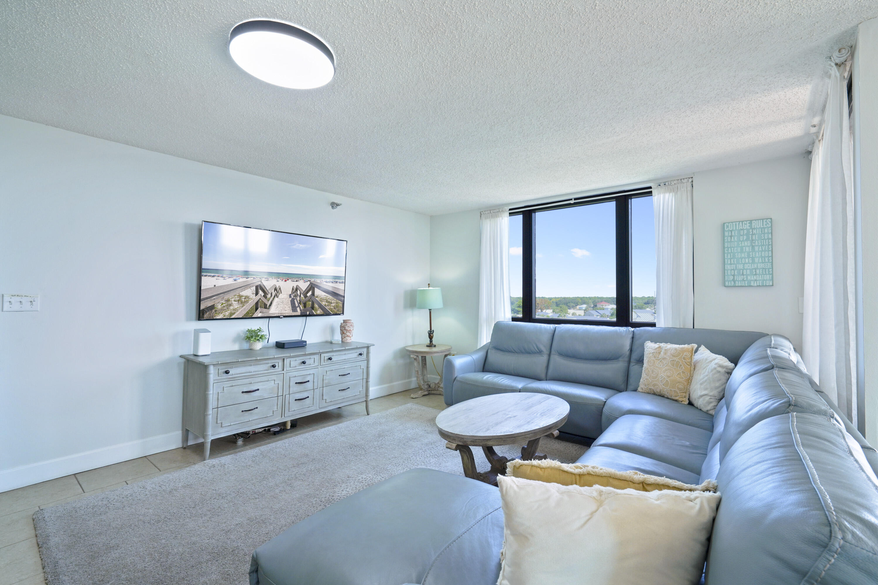 Living room with blue sectional sofa, oval coffee table, and large windows overlooking rooftops and trees.