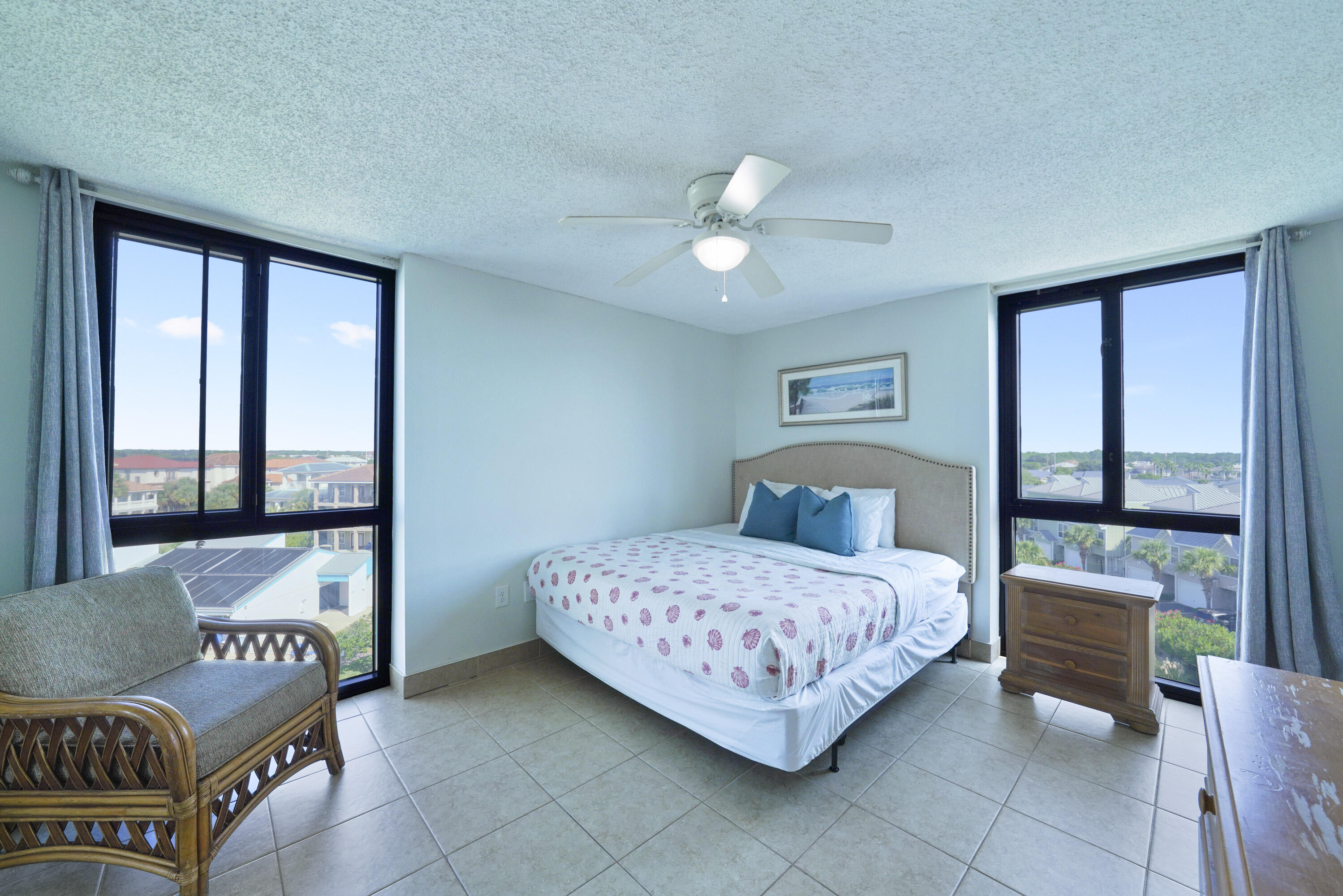 Bedroom with a queen bed, wicker chair, and two large windows overlooking nearby homes.