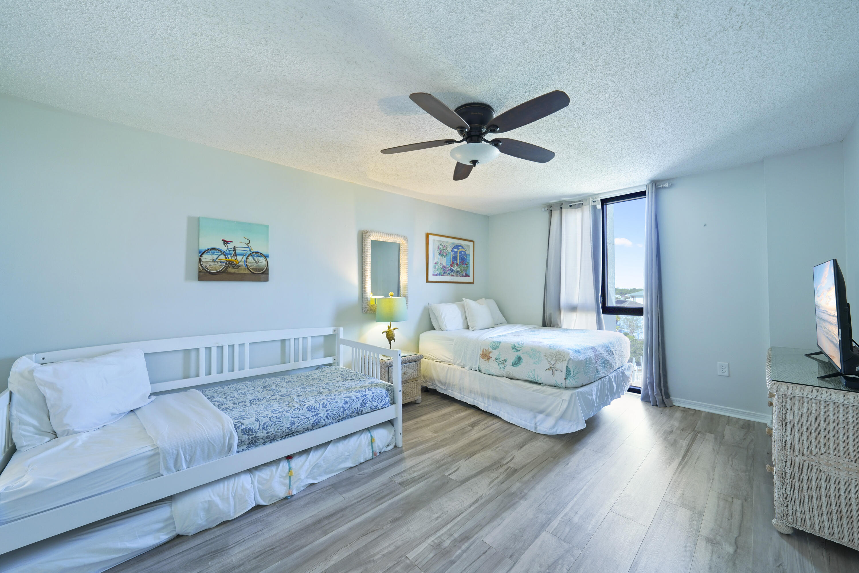 Furnished bedroom with a white daybed, queen bed, coastal artwork, and tall window.