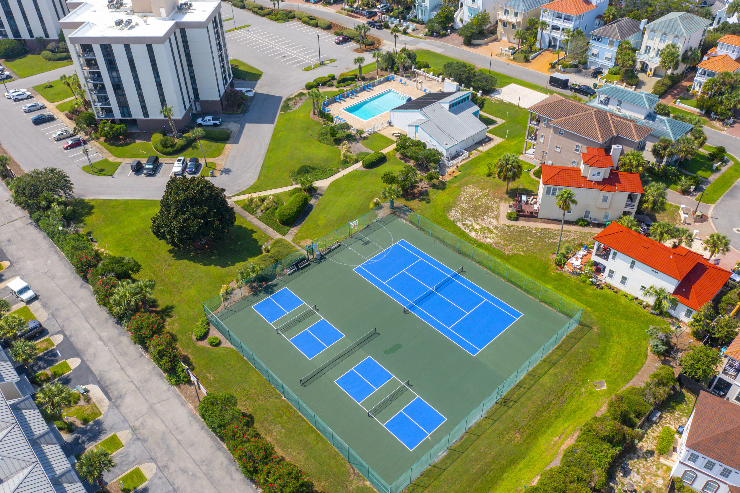 Aerial view of condominium grounds with blue courts, a swimming pool, and landscaped lawns.
