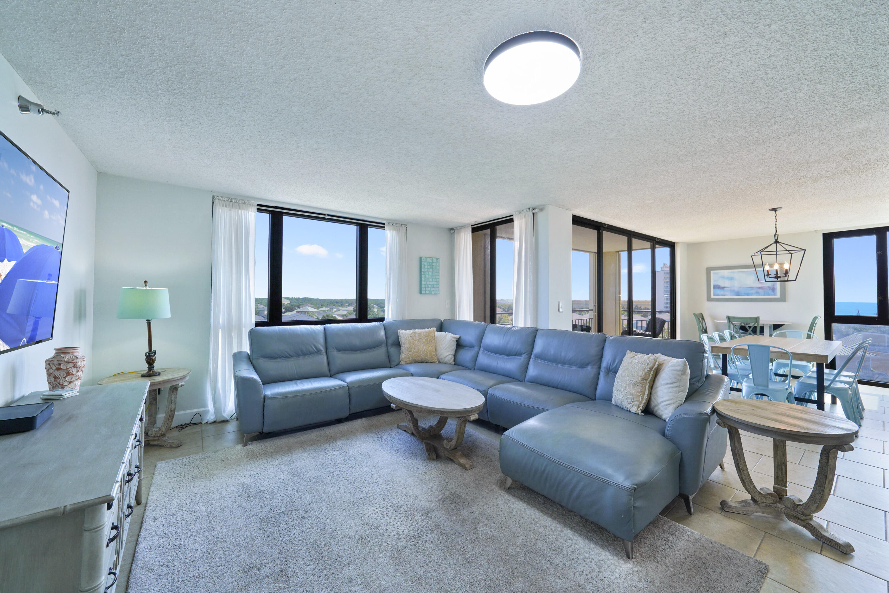 Furnished condo living and dining area with blue sectional and floor-to-ceiling windows overlooking the water.