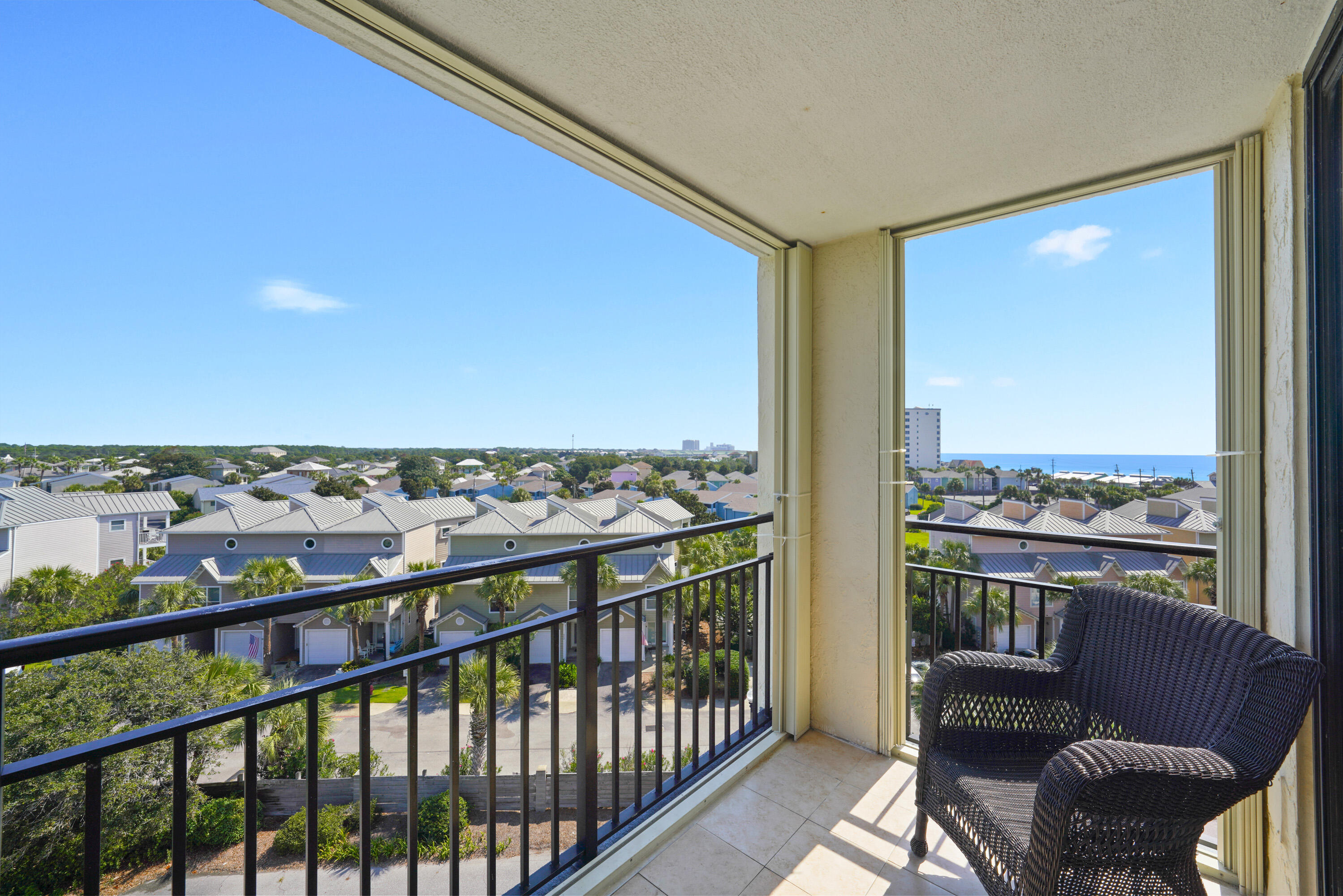 Condo balcony with wicker chairs overlooking neighborhood rooftops and distant water.