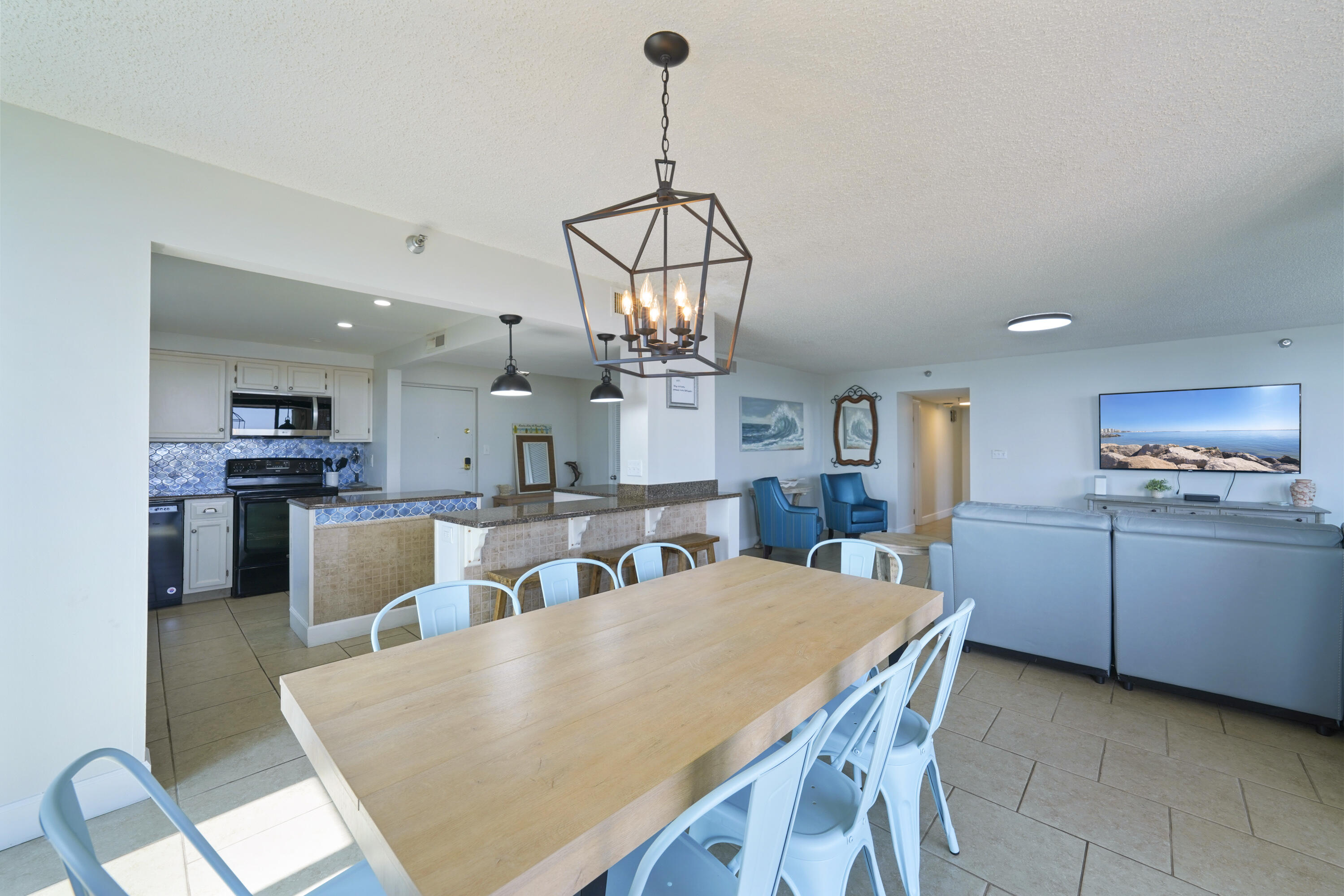 Open-plan condo kitchen and dining area with blue backsplash, large wood table, and geometric pendant light.