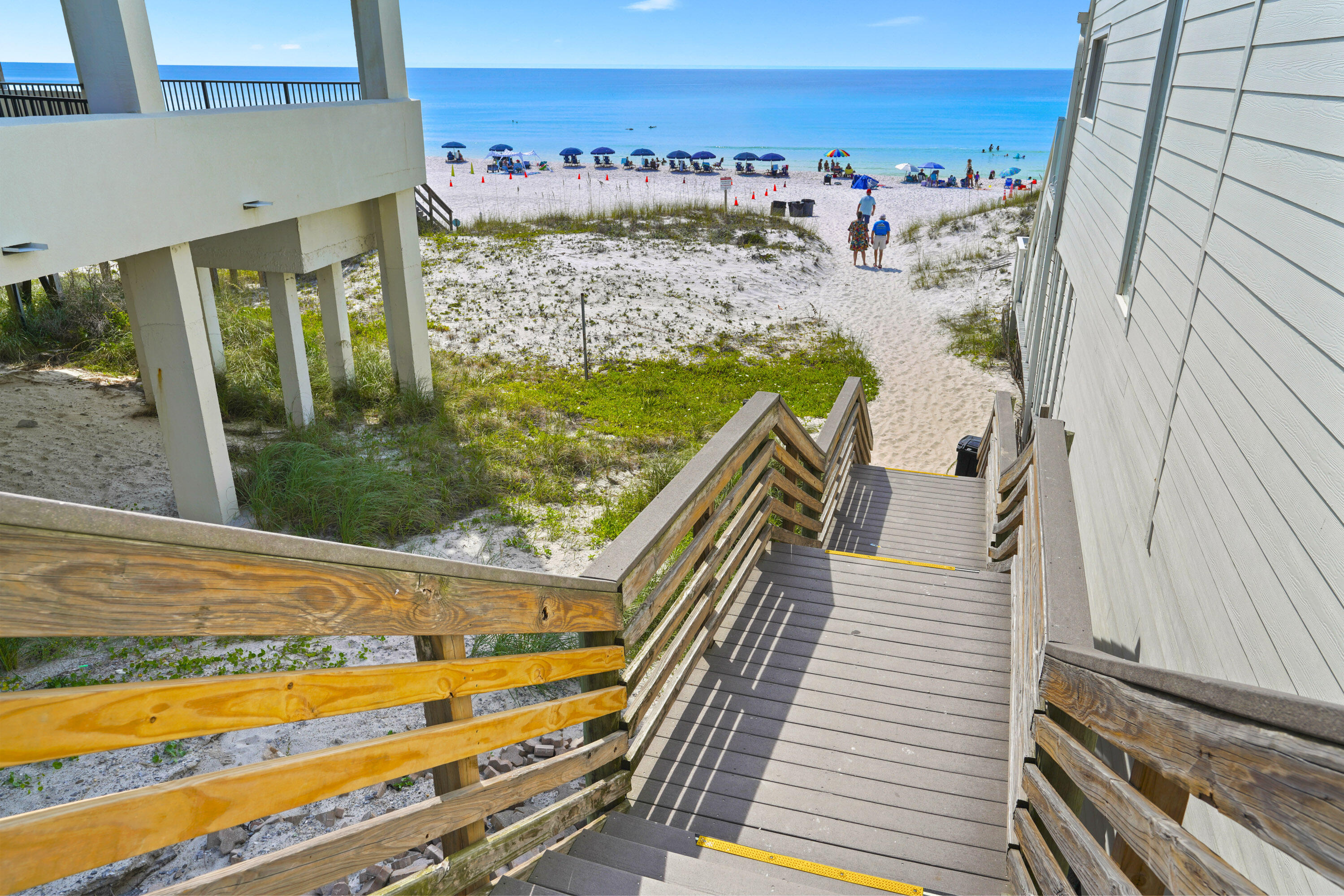 Beach access stairs lead through dunes to a sandy beach with umbrellas and blue Gulf water.