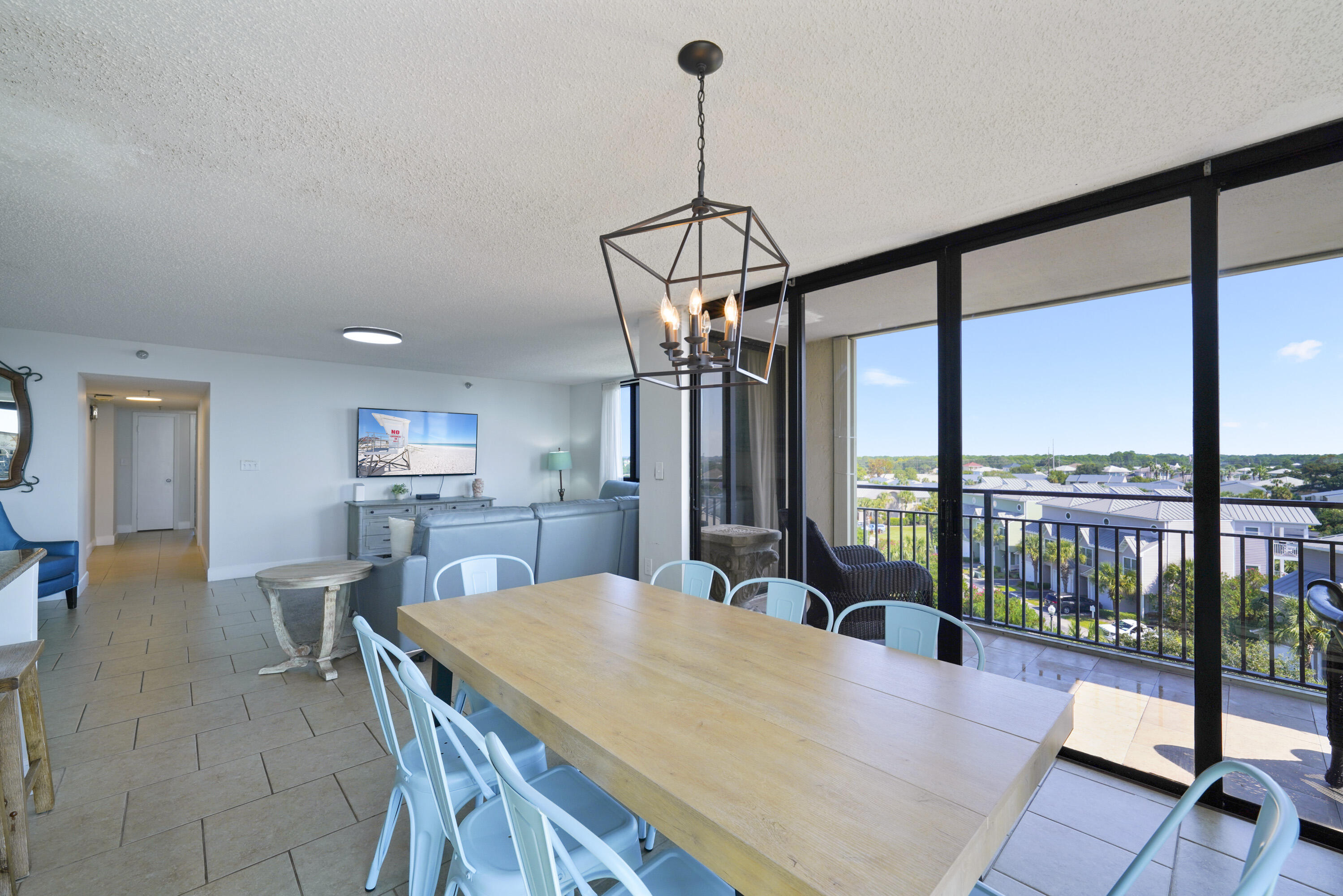 Condo dining area with a long wood table and blue chairs beside glass doors opening to a sunny balcony view.
