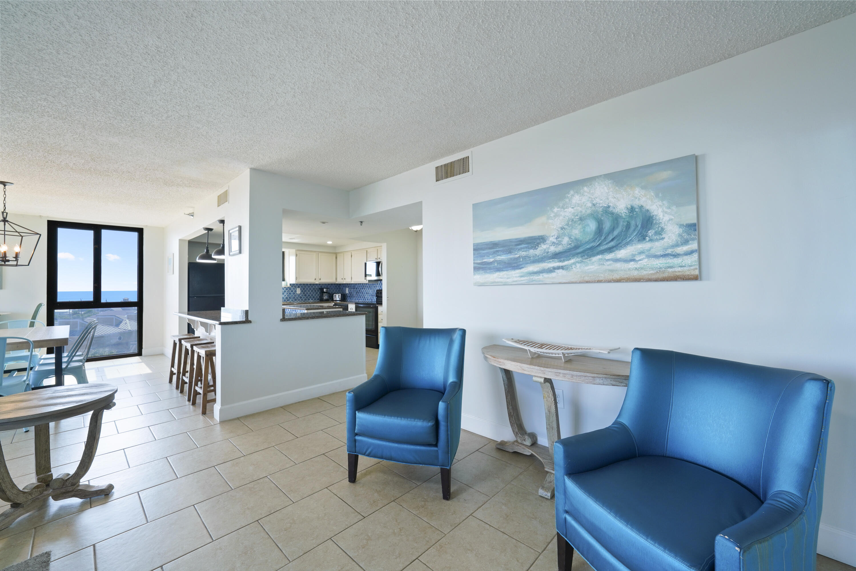 Open-plan living area with blue armchairs, a tiled kitchen, and ocean views through large windows.