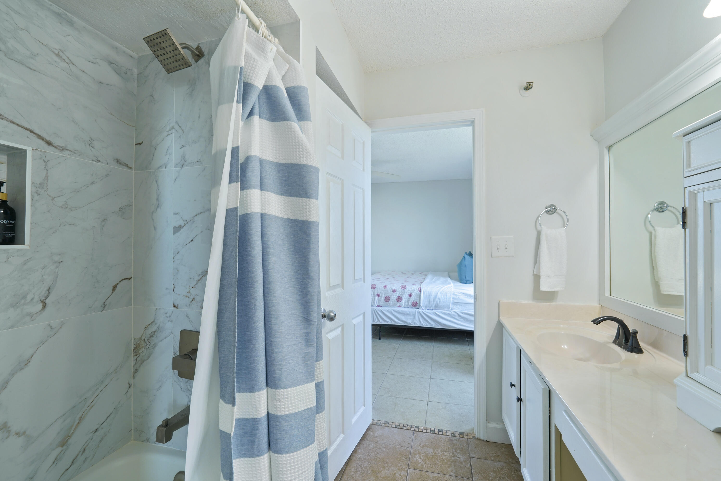 Bathroom with marble-look shower walls, a blue striped curtain, and a vanity beside an open bedroom doorway.