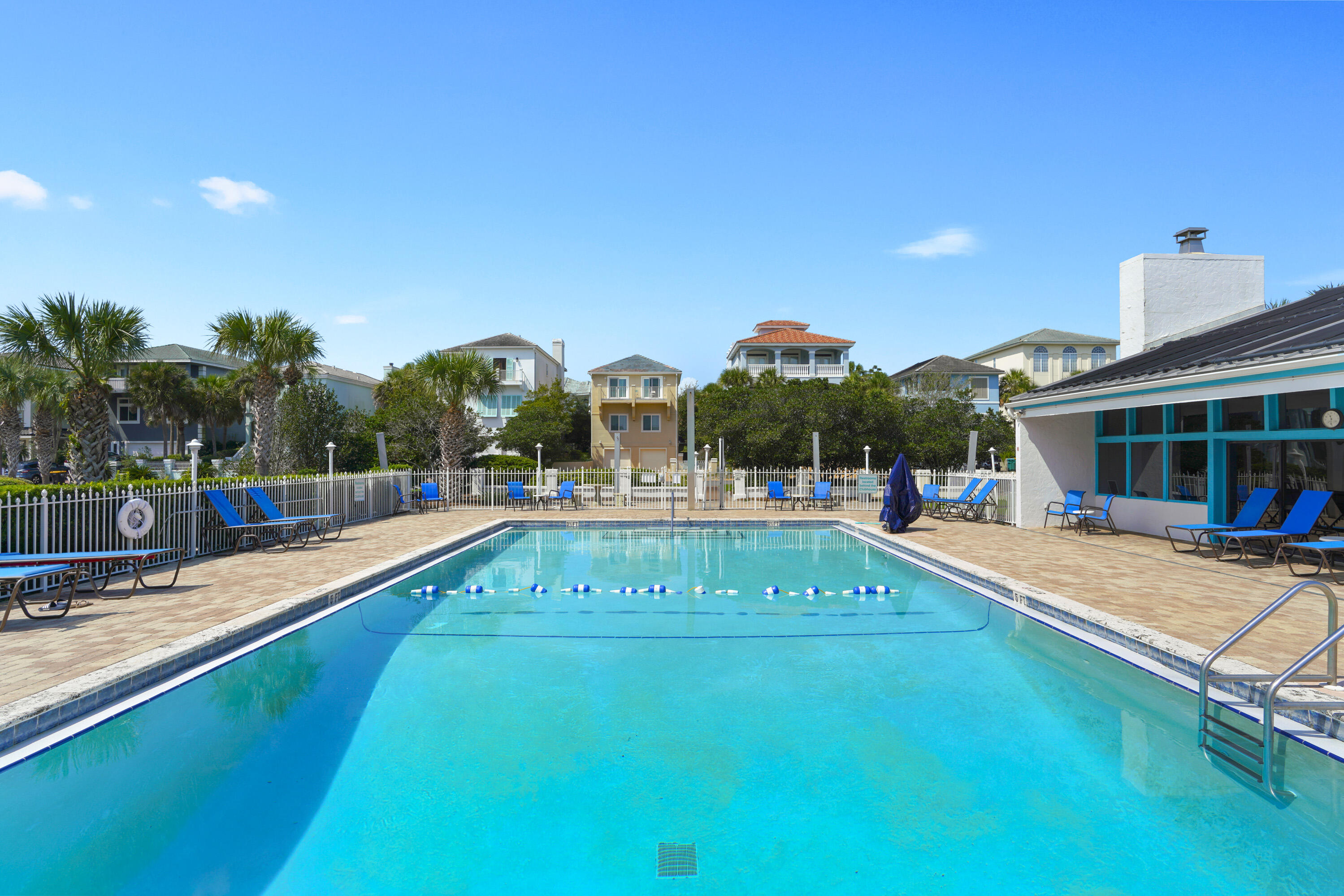 Outdoor swimming pool with a fenced brick deck and blue lounge chairs.