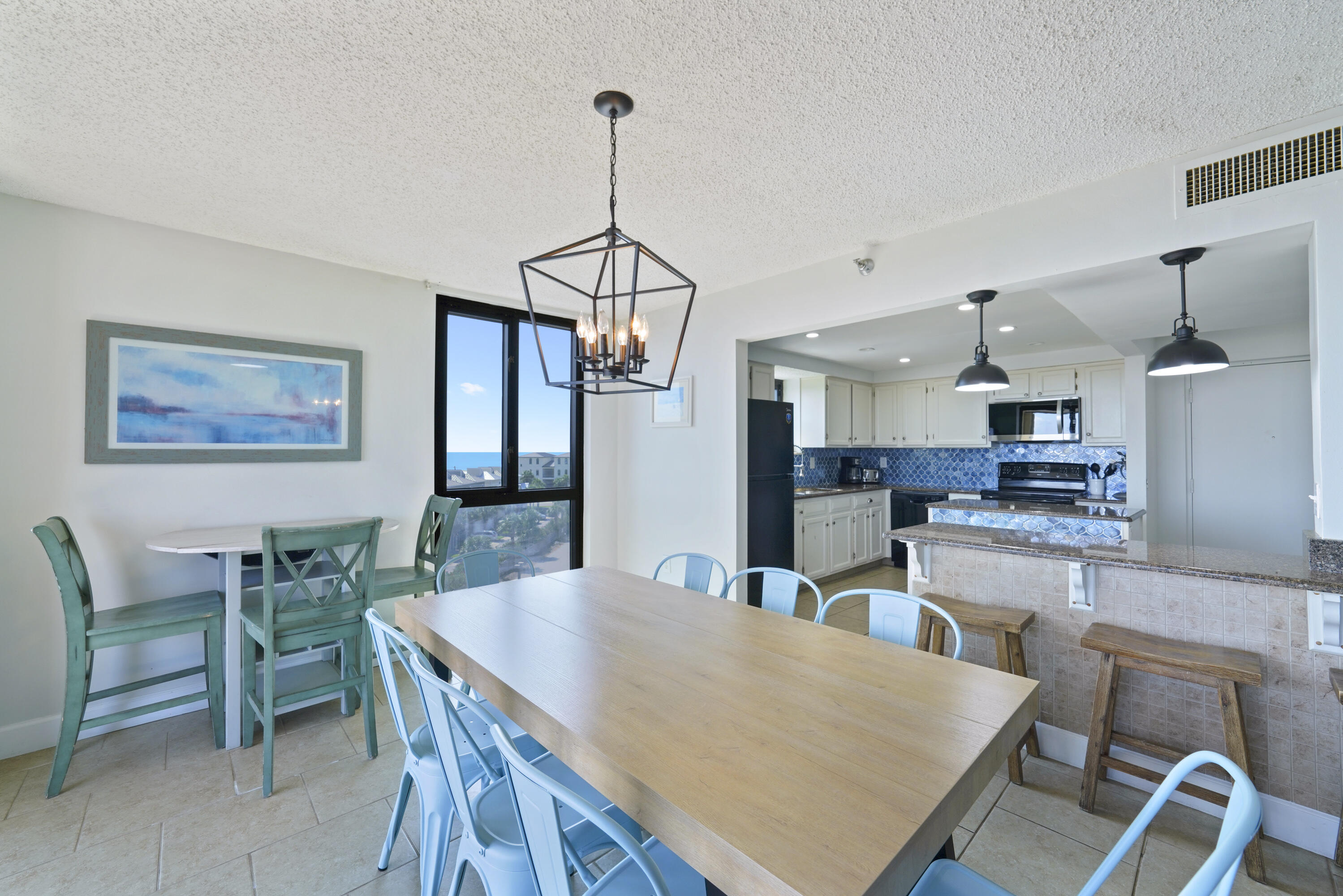 Dining area and kitchen with blue tile backsplash, turquoise chairs, and a glimpse of the sea through the window.