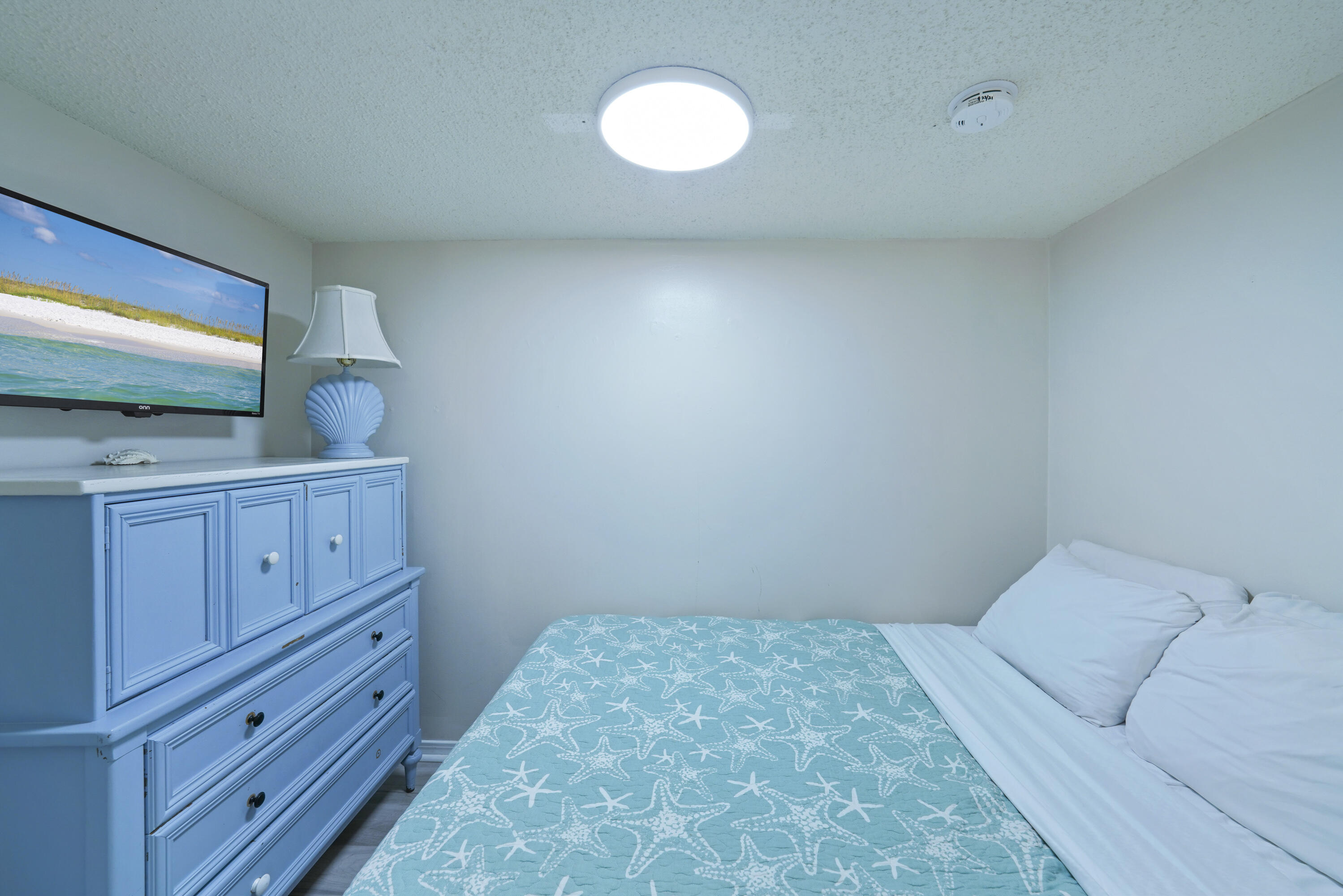 Bedroom with a turquoise starfish quilt, blue dresser, and television showing a beach.