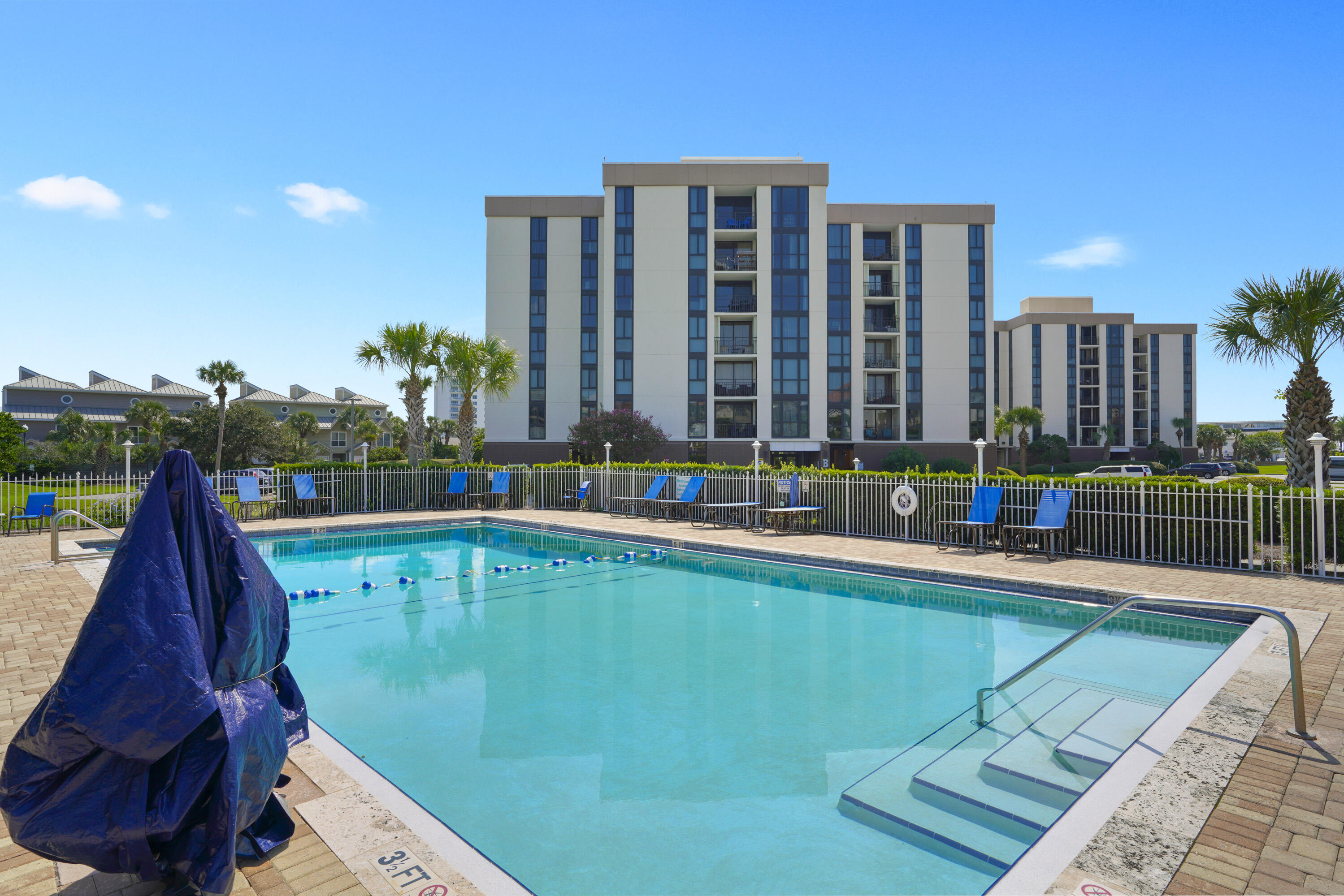 Condominium pool with a shallow-entry area, blue lounge chairs, and palm trees.