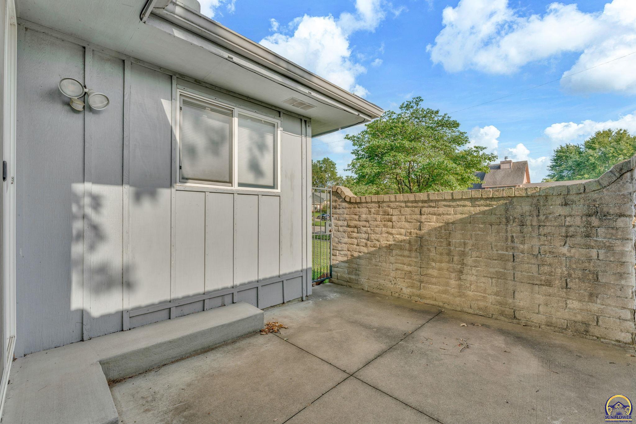 Concrete patio beside gray siding, enclosed by a tan block privacy wall and metal gate.