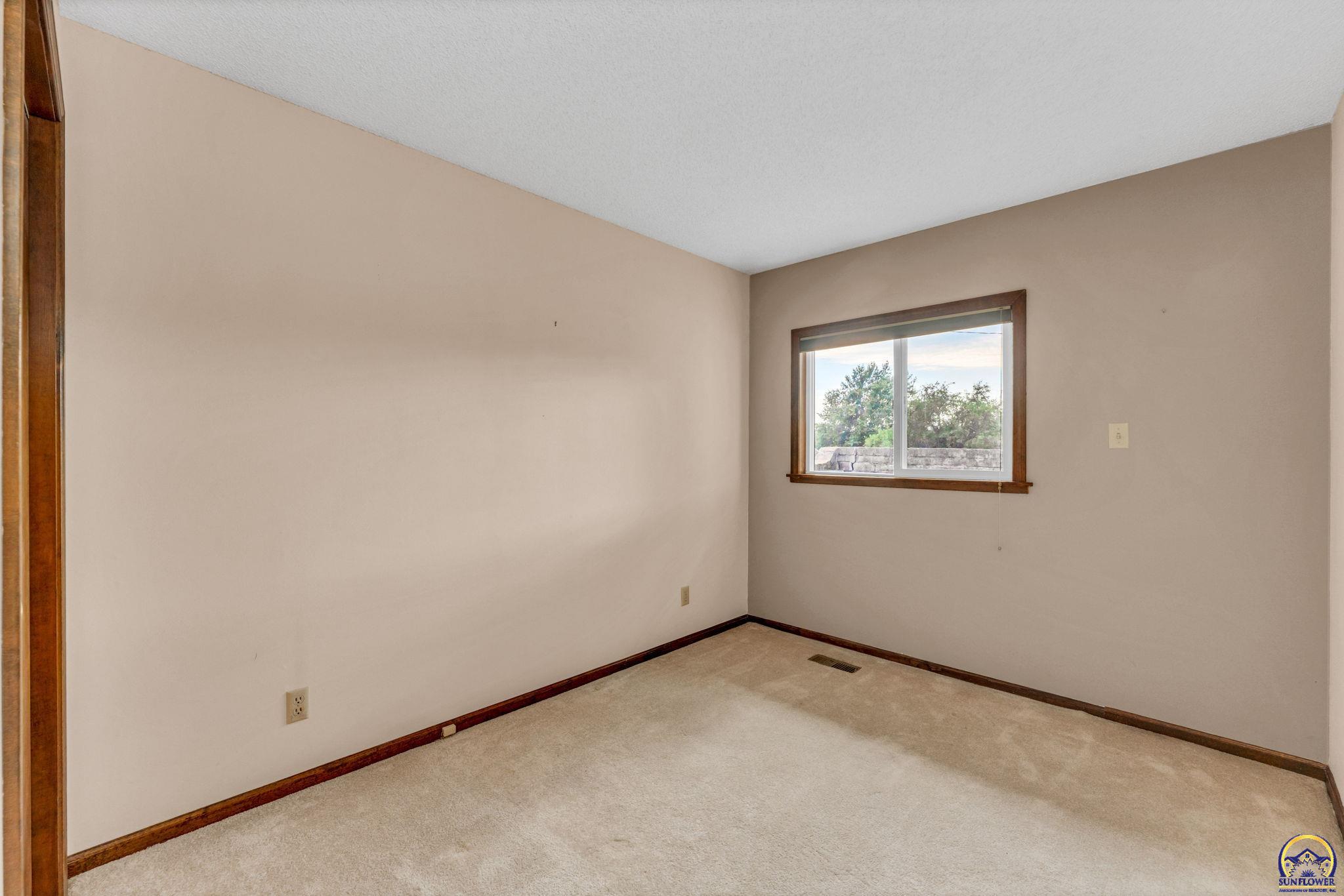 Empty carpeted room with dark wood trim and a window overlooking trees.